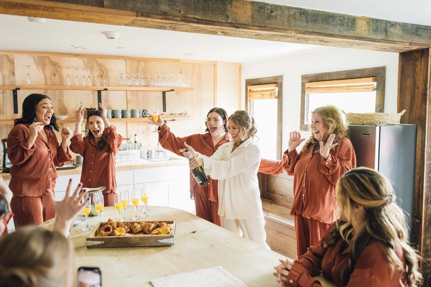 Bridesmaids in matching pajamas celebrating with champagne in the bright kitchen of the bridal suite at The Barn at Villa Venezia.