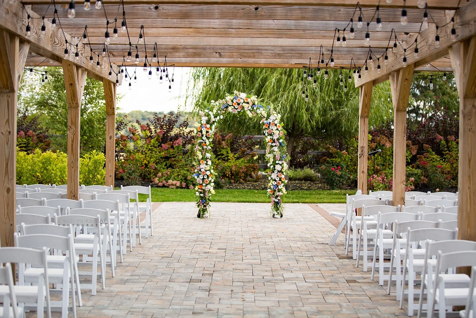 Outdoor wedding ceremony setup under a wooden pergola with white chairs and a colorful floral arch for a summer wedding in New York