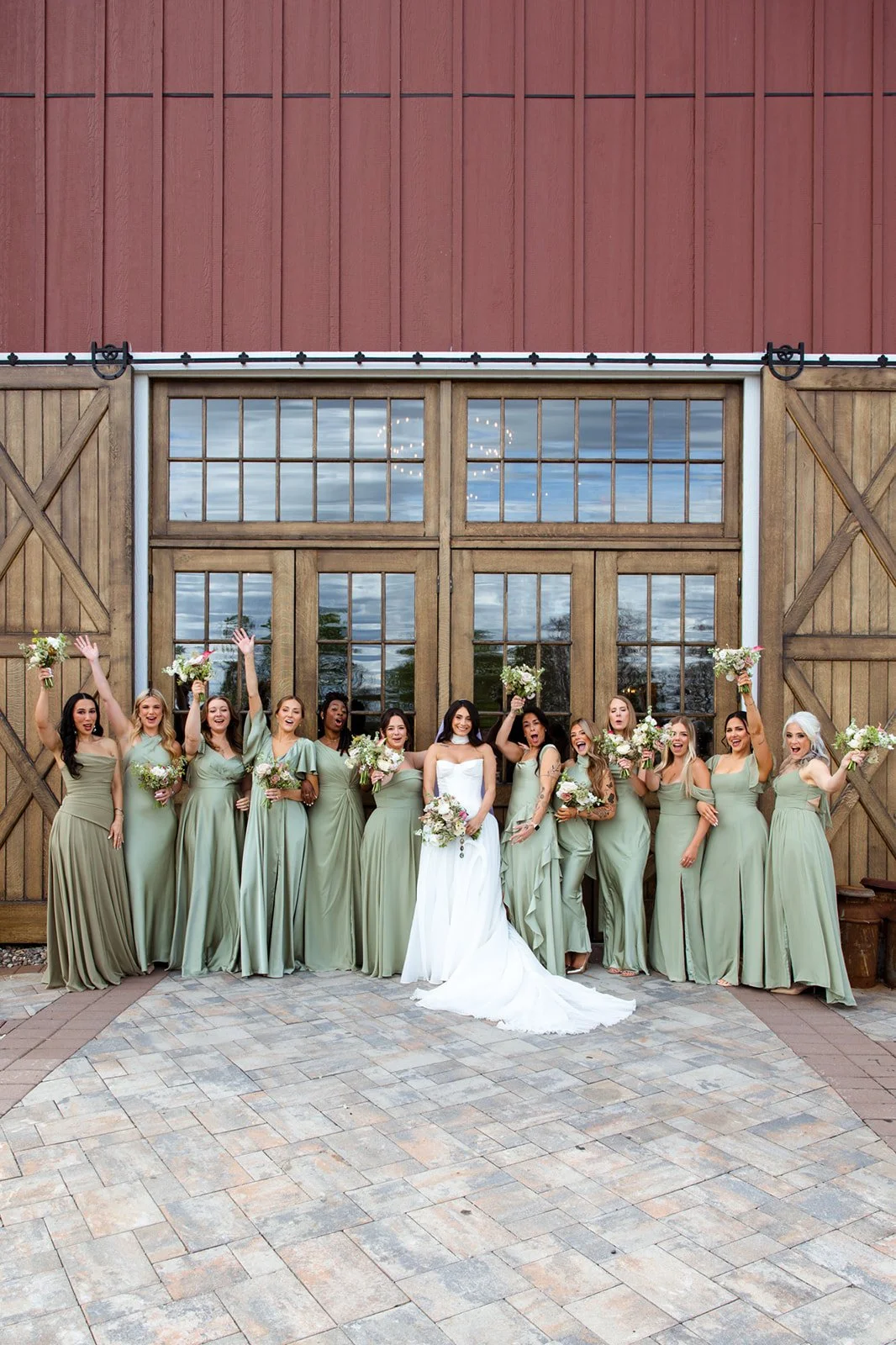 Large bridesmaid group cheering in mismatched sage green dresses against the towering wooden barn doors.