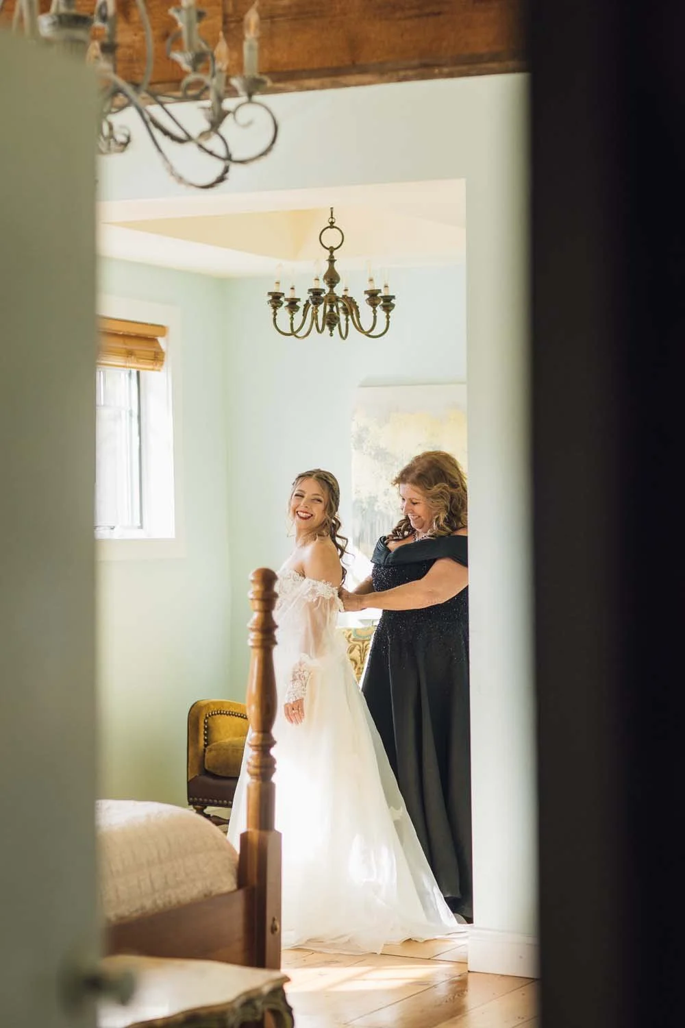A mother smiling as she buttons the back of her daughter's wedding gown in a bright, sunlit bridal dressing room.
