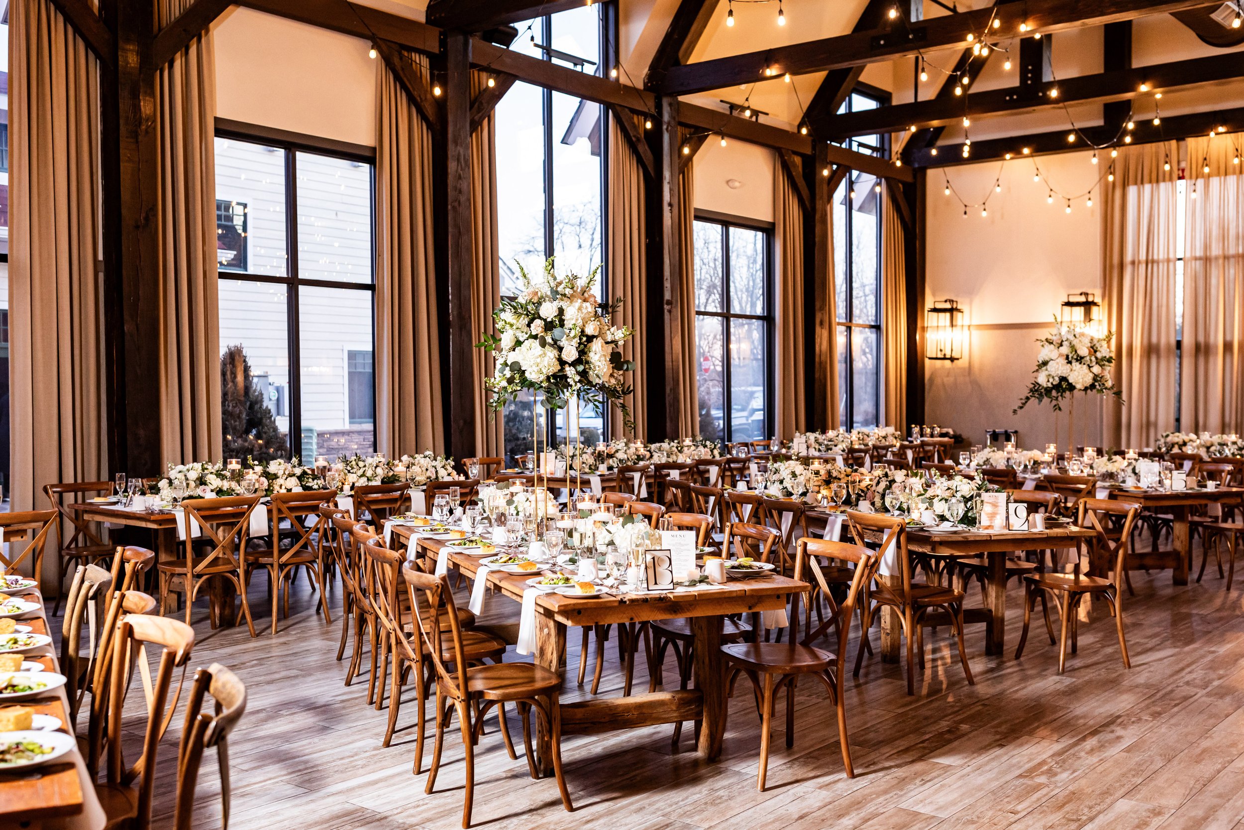 Indoor wedding reception at The Barn at Villa Venezia featuring exposed wooden beams, hanging string lights, rustic farm tables with cross-back chairs, and tall white floral centerpieces.