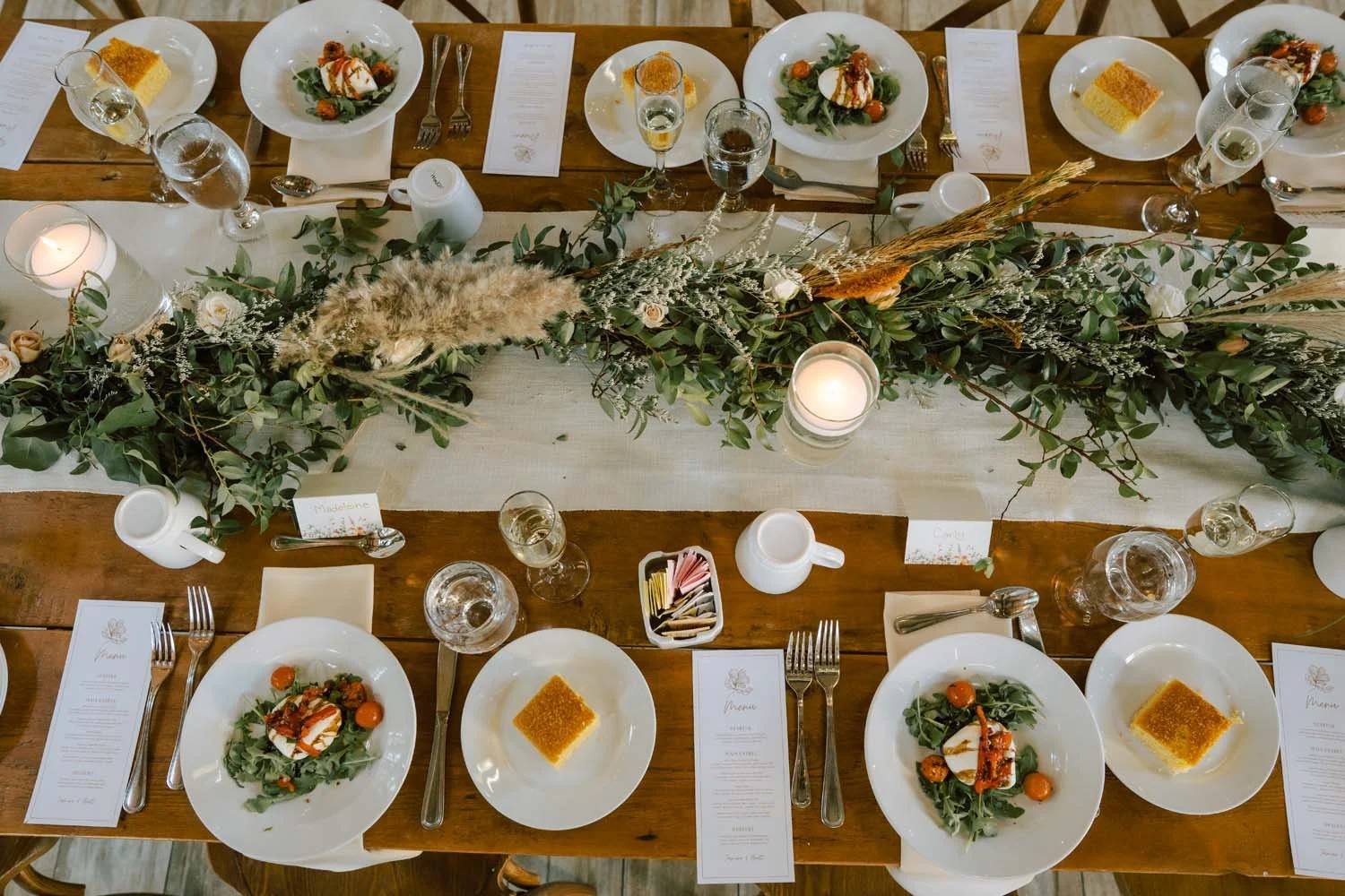 Overhead view of wooden farm table with eucalyptus runner, floating candles, and plated burrata salad