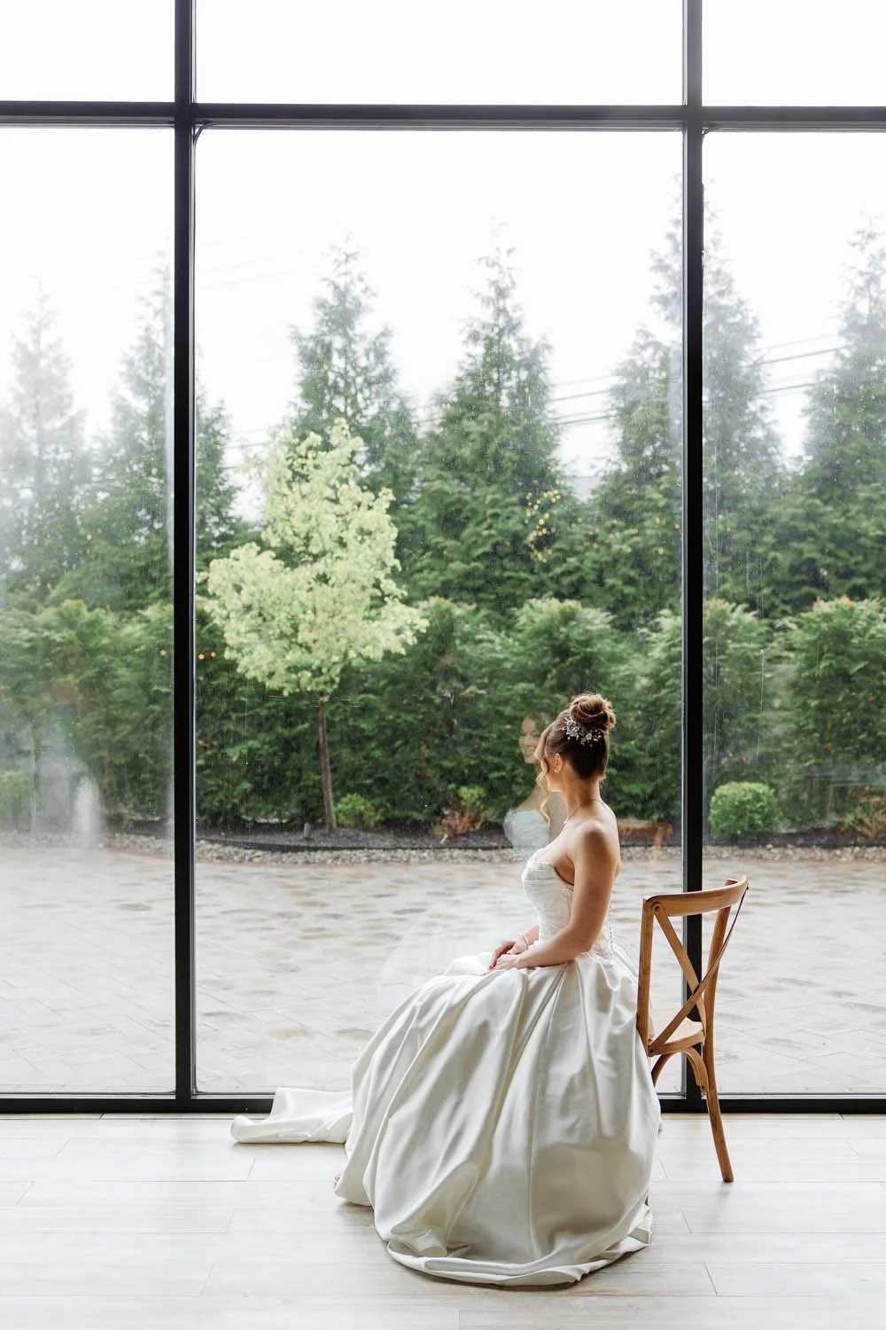 Silhouette-style portrait of a bride sitting on a wooden crossback chair looking out the giant floor-to-ceiling
