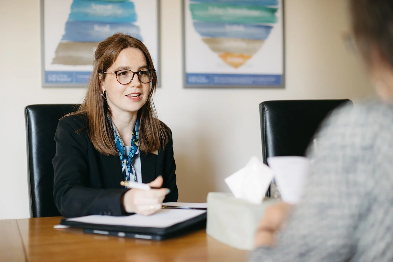 A woman with brown hair, wearing glasses, a black blazer, and a blue patterned scarf, is sitting at a wooden table during a business meeting. She is holding a pen, with a folder in front of her, and is engaged in conversation with a person whose back is to the camera. Two colorful charts are visible on the wall behind her.