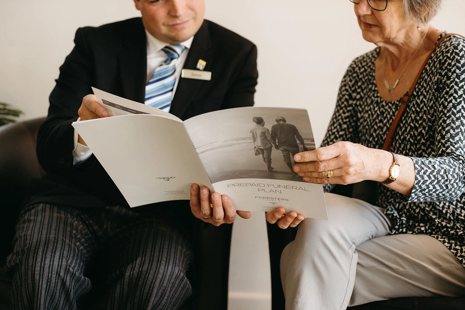A man in a suit and woman in patterned blouse looking at a booklet titled 'Prepaid Funeral Plan' while sitting on a couch.