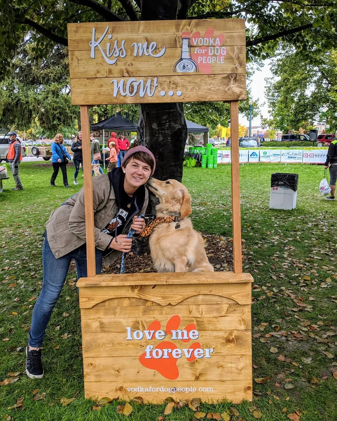 A woman with a dog under a wooden signframe at an outdoor event. The sign reads 'Kiss me now...' and 'love me forever' with a paw print and the website vodkafordogpeople.com. The woman is smiling and holding the dog, which is licking her face. In the background, other people are walking and enjoying the event.