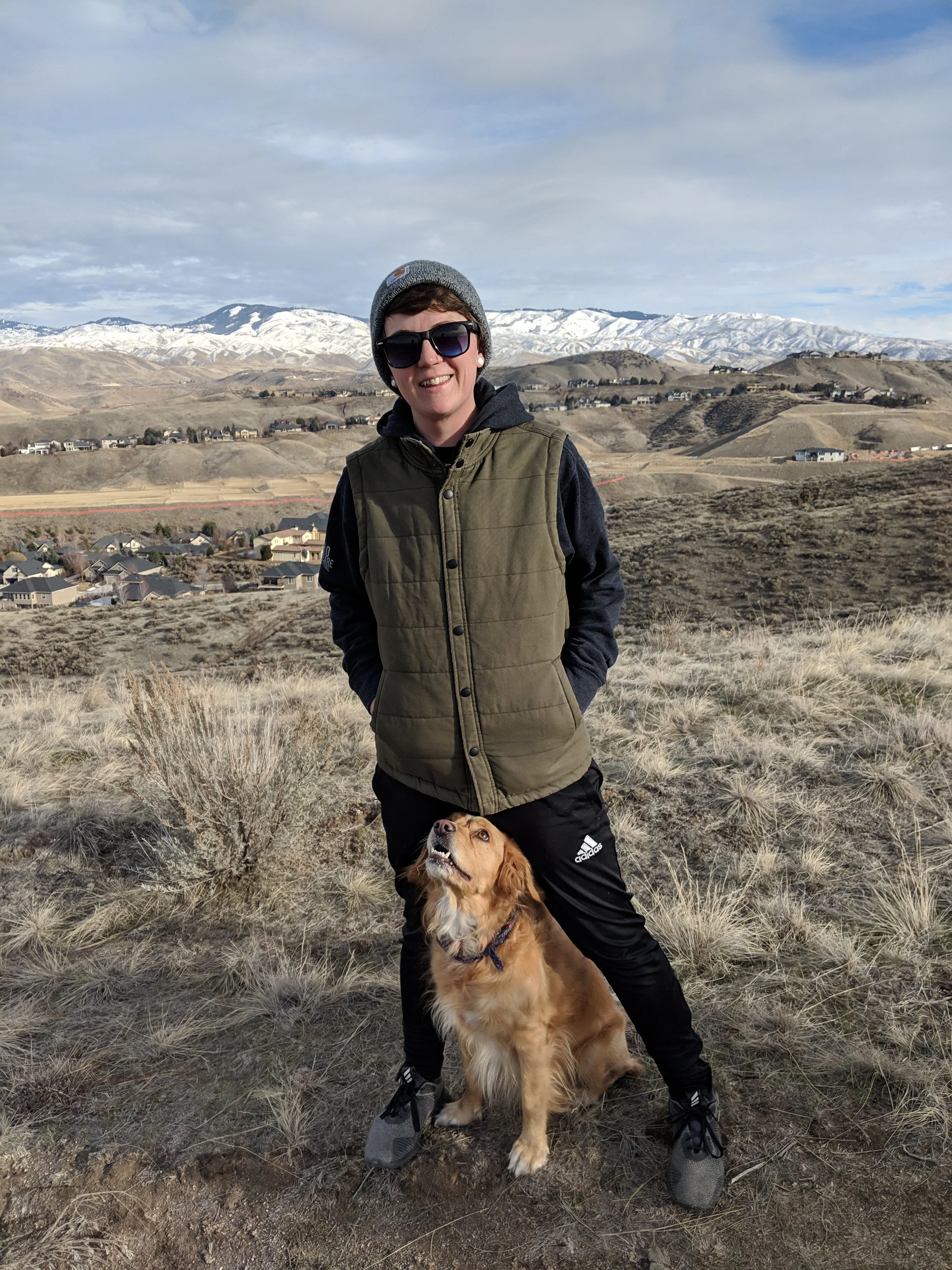 A Boise woman in sunglasses and a beanie standing outdoors with her dog in a dry, hilly landscape with mountains in the background.