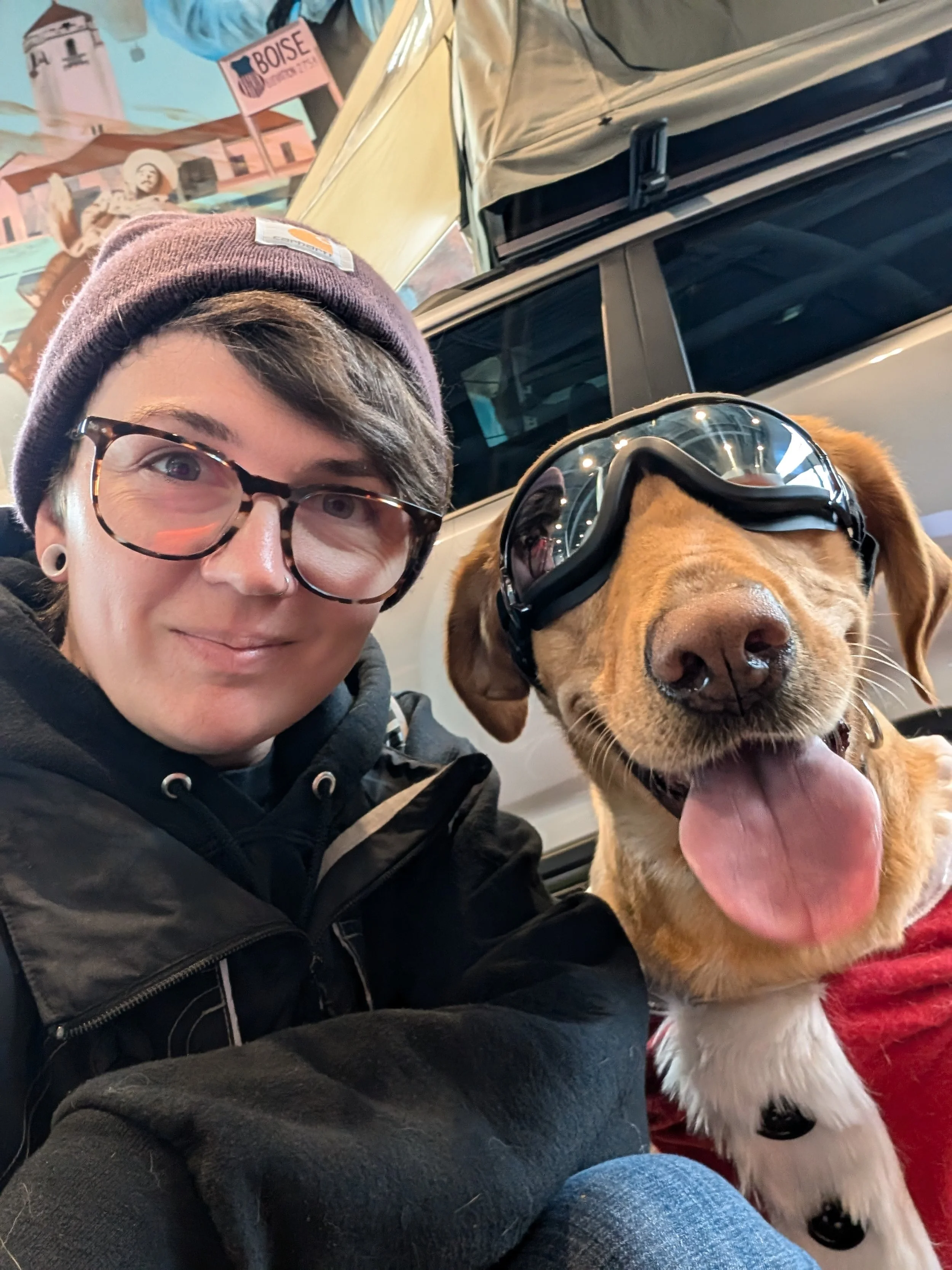 A person wearing glasses, a burgundy beanie, and a black hoodie taking a selfie with a dog that has goggles on its eyes, its tongue out, and wearing a red and white sweater. They are indoors with a vehicle and mural in the background. Boise.