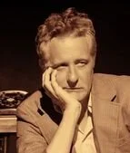 A young man with curly hair resting his head on his hand, wearing a blazer, in a vintage-style setting.