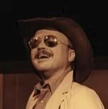 A man wearing a wide-brimmed hat and sunglasses, smiling and dressed in a light-colored jacket.