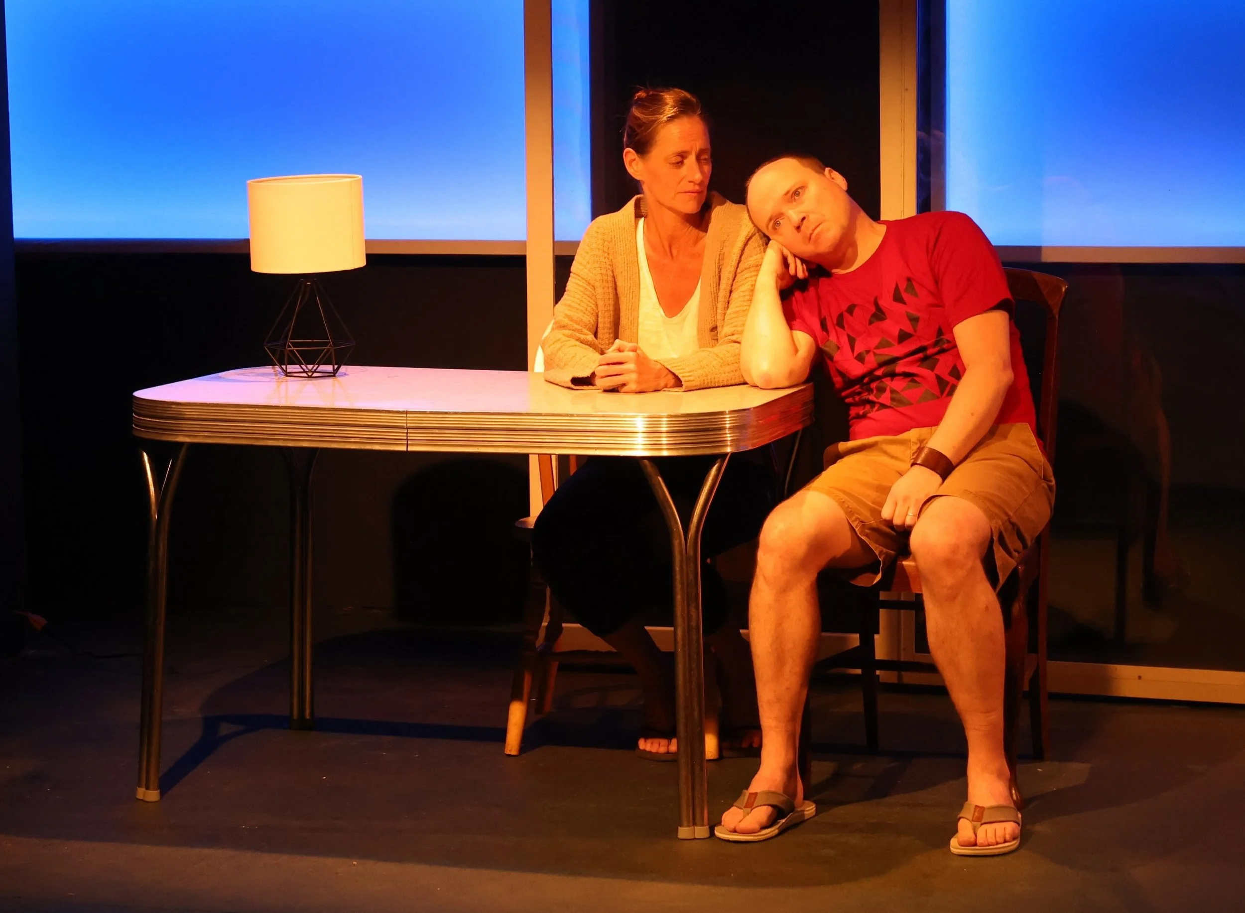 A woman and a man sit at a table with a lamp on it, with blue and black background, and the woman appears to be comforting or having a serious conversation with the man who is resting his head on his hand.