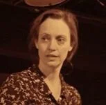 A woman with short brown hair wearing a patterned top, looking to her left in a dimly lit environment.