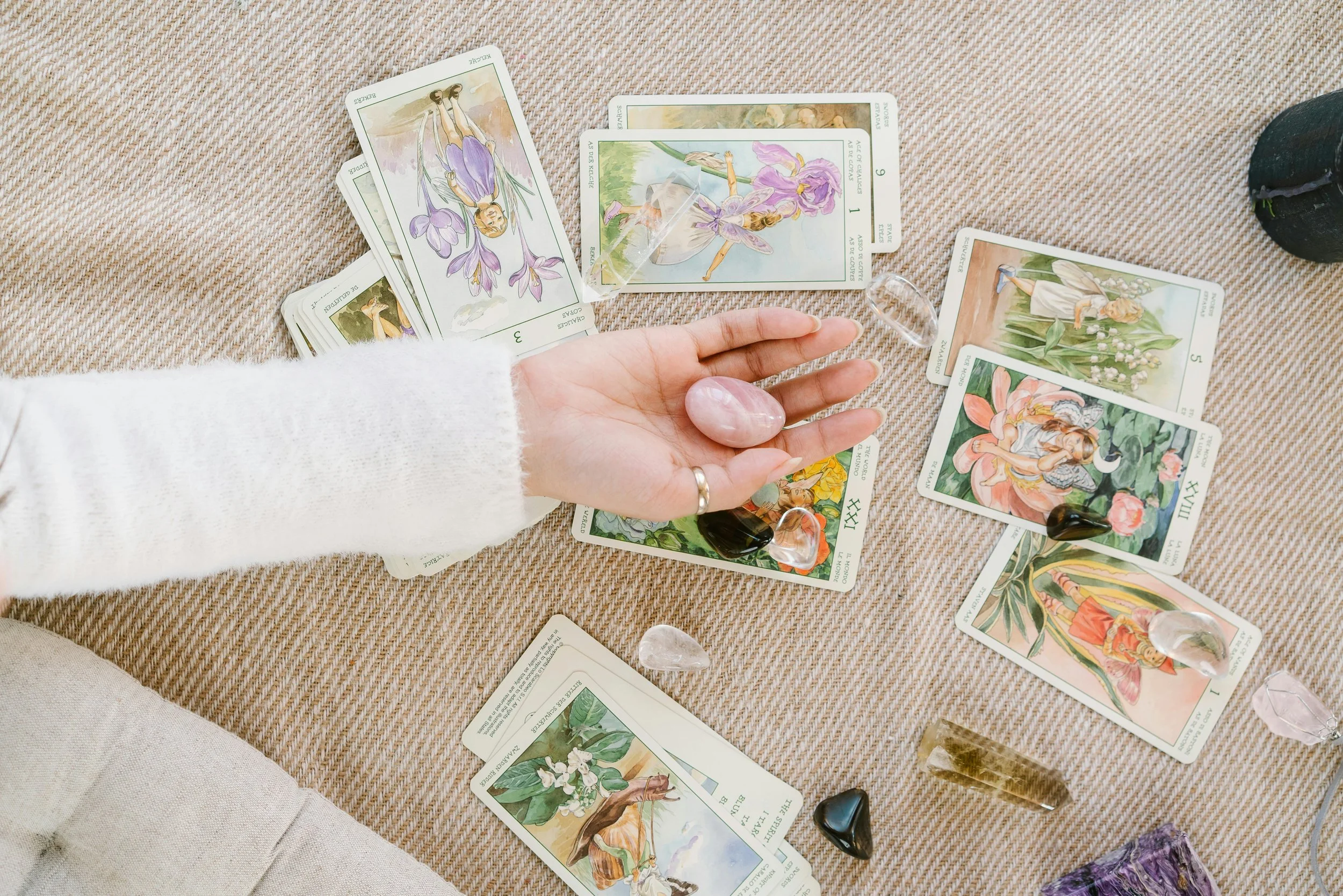 Person with a white sleeve and ring on finger arranging tarot cards and healing crystals on a beige fabric surface.