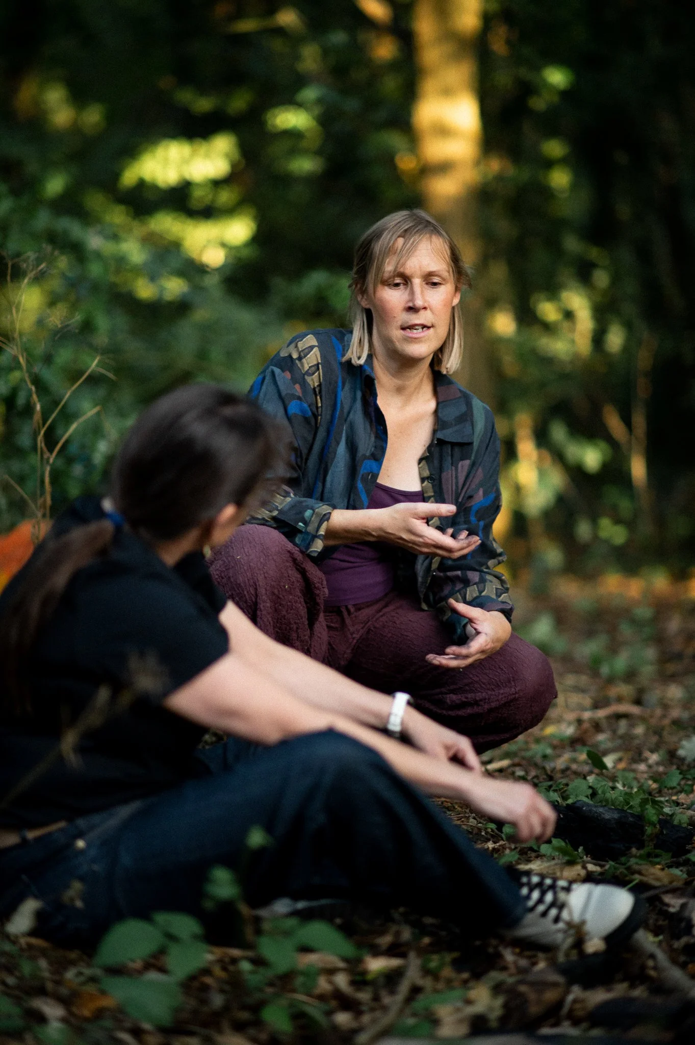 Two women sitting on the ground in a wooded area, engaged in conversation, with one woman gesturing as she speaks.