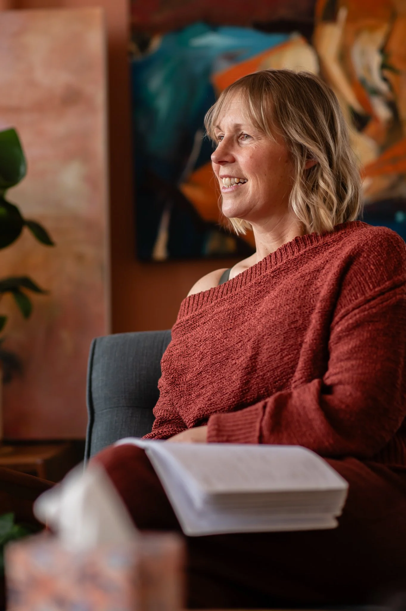 A woman with blonde hair smiling while seated indoors with abstract artwork in the background and an open book on her lap.