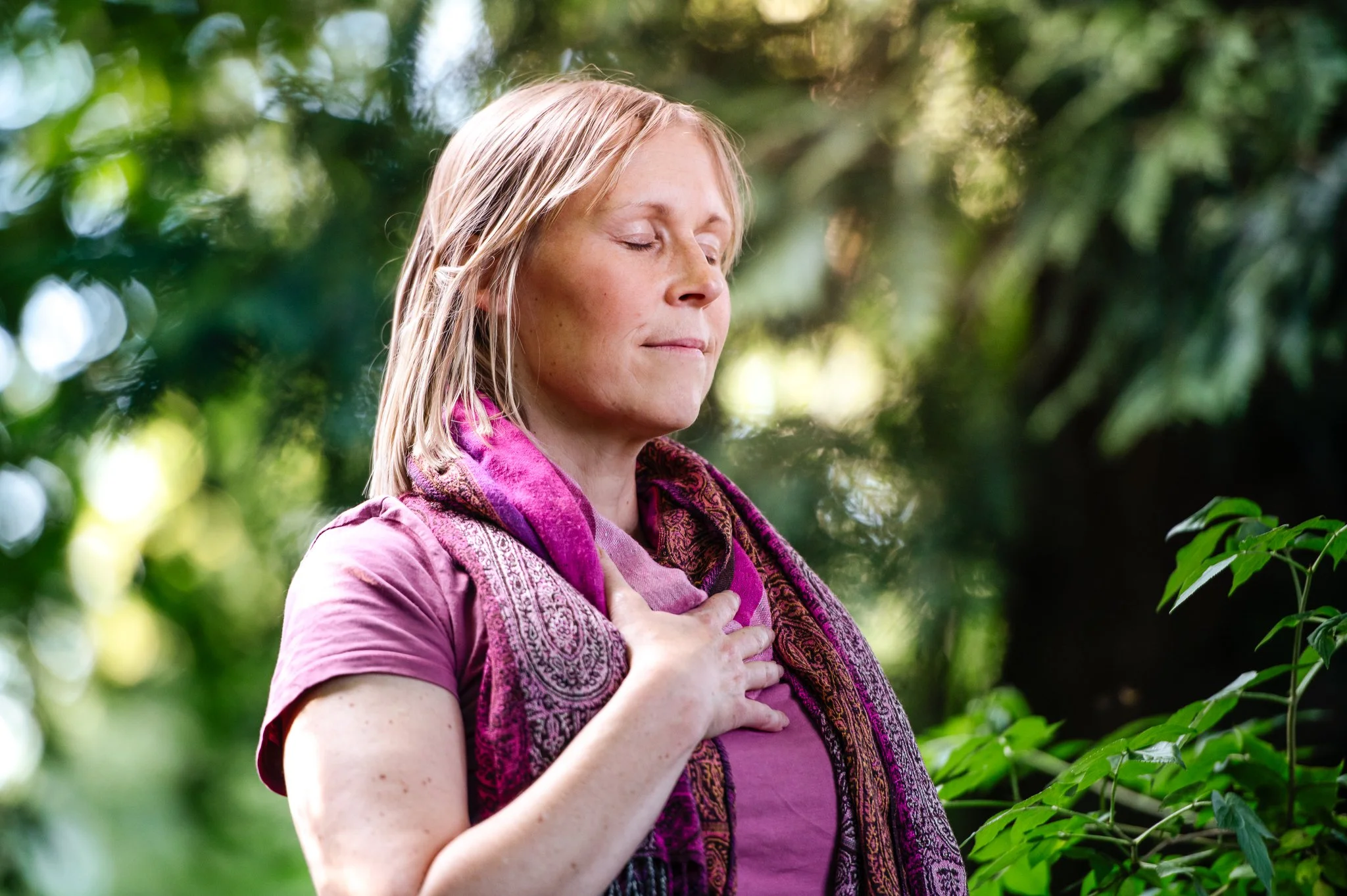 A woman with blonde hair, eyes closed, standing outdoors with greenery, holding her hand on her chest as if meditating or practicing mindfulness.