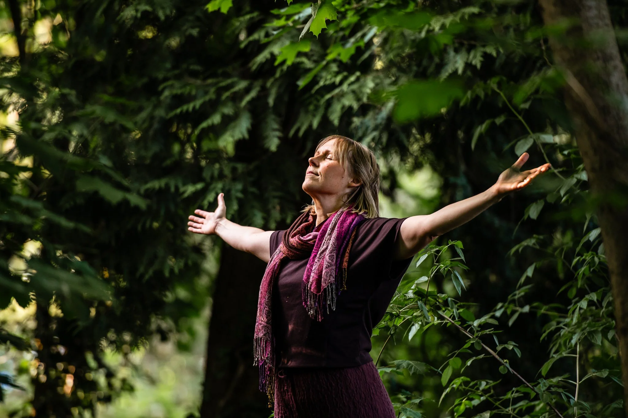 A woman standing in a lush green forest with arms outstretched and eyes closed, appearing to enjoy the moment and connect with nature.