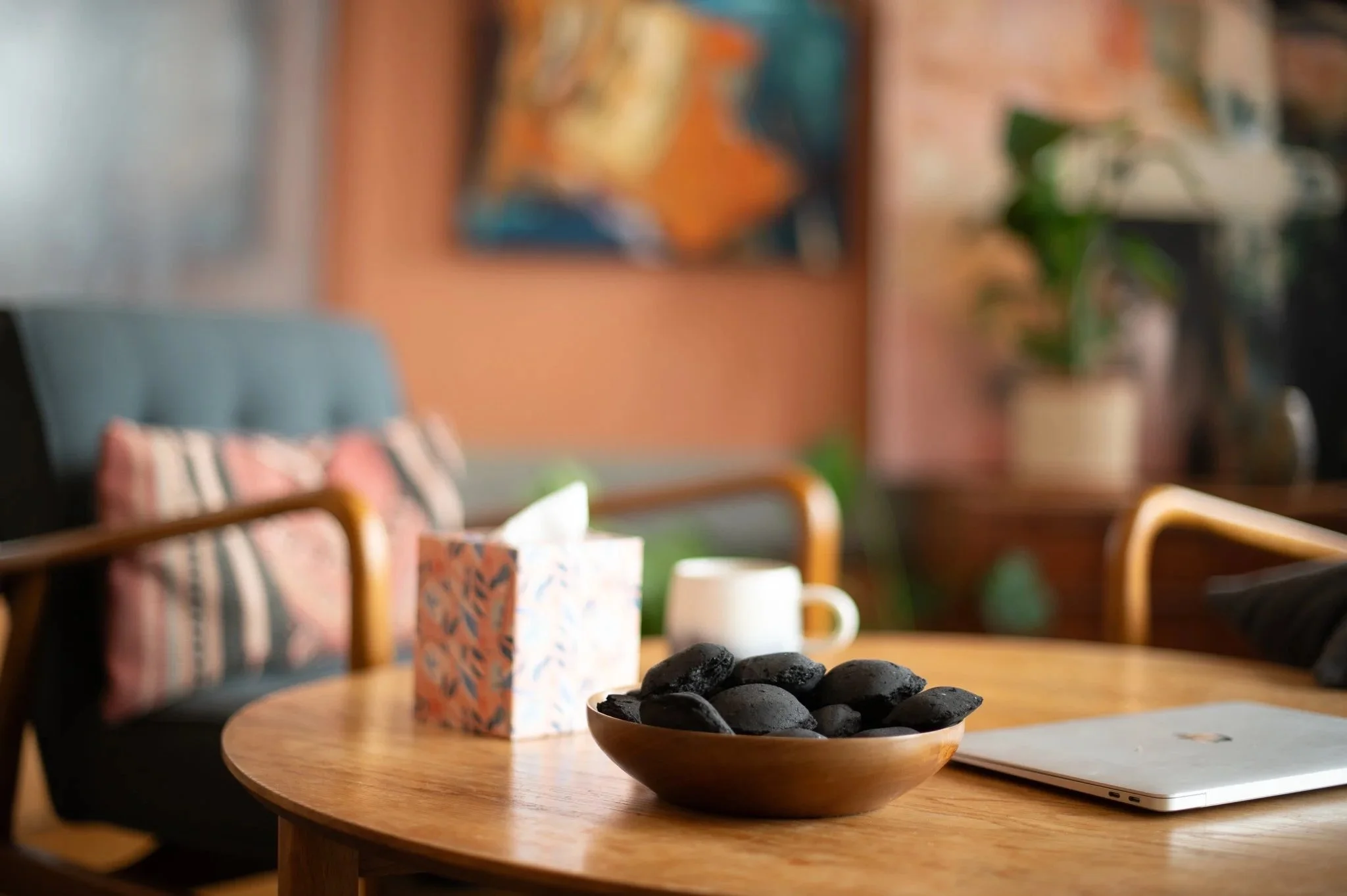 A wooden table with a bowl of black embers, a colorful tissue box, a white coffee mug, and a closed laptop in a cozy living room with colourful artwork and plants in the background.