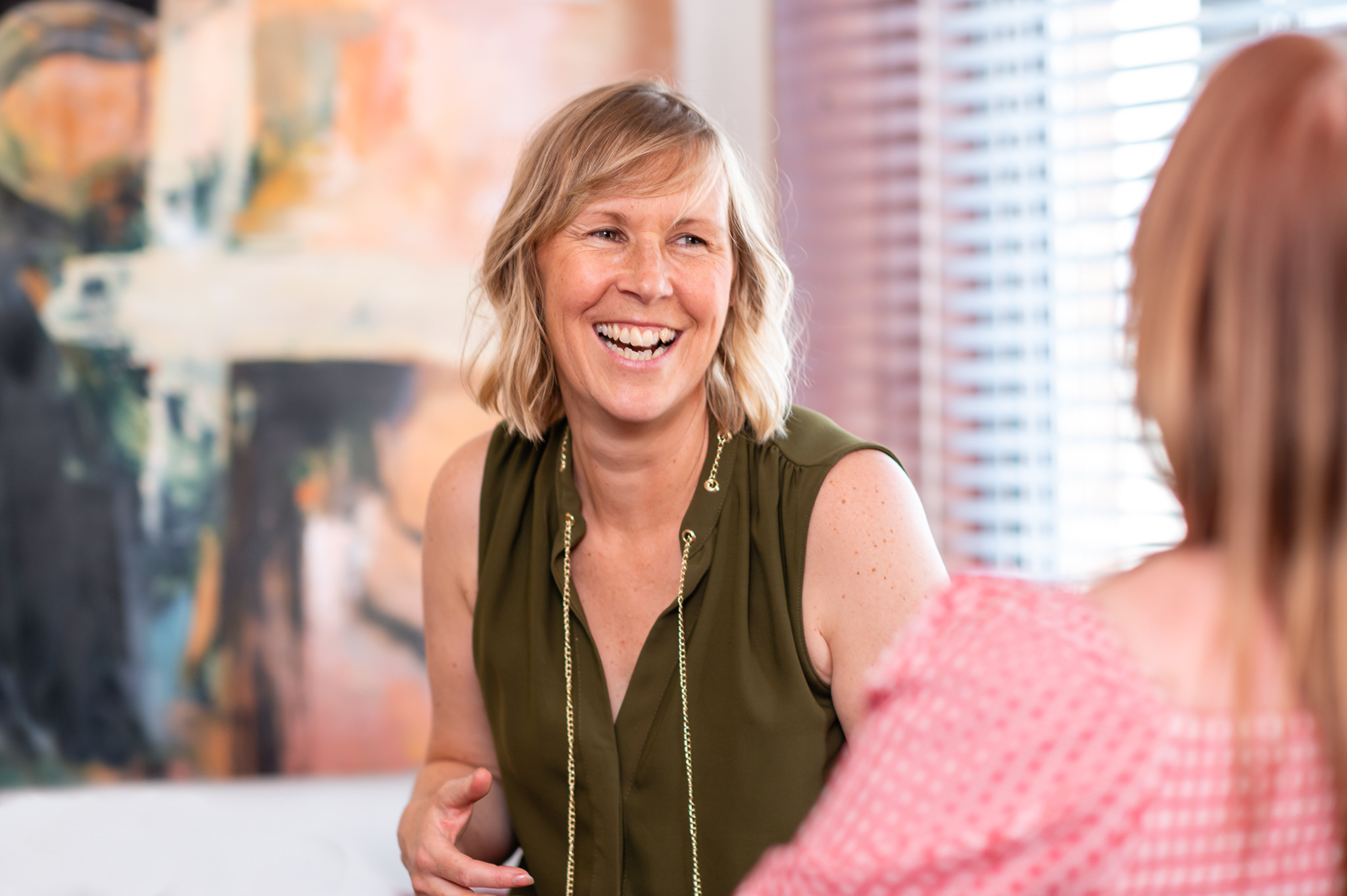 Julia Hamer smiling and engaging in conversation with another woman with abstract artwork and window blinds.