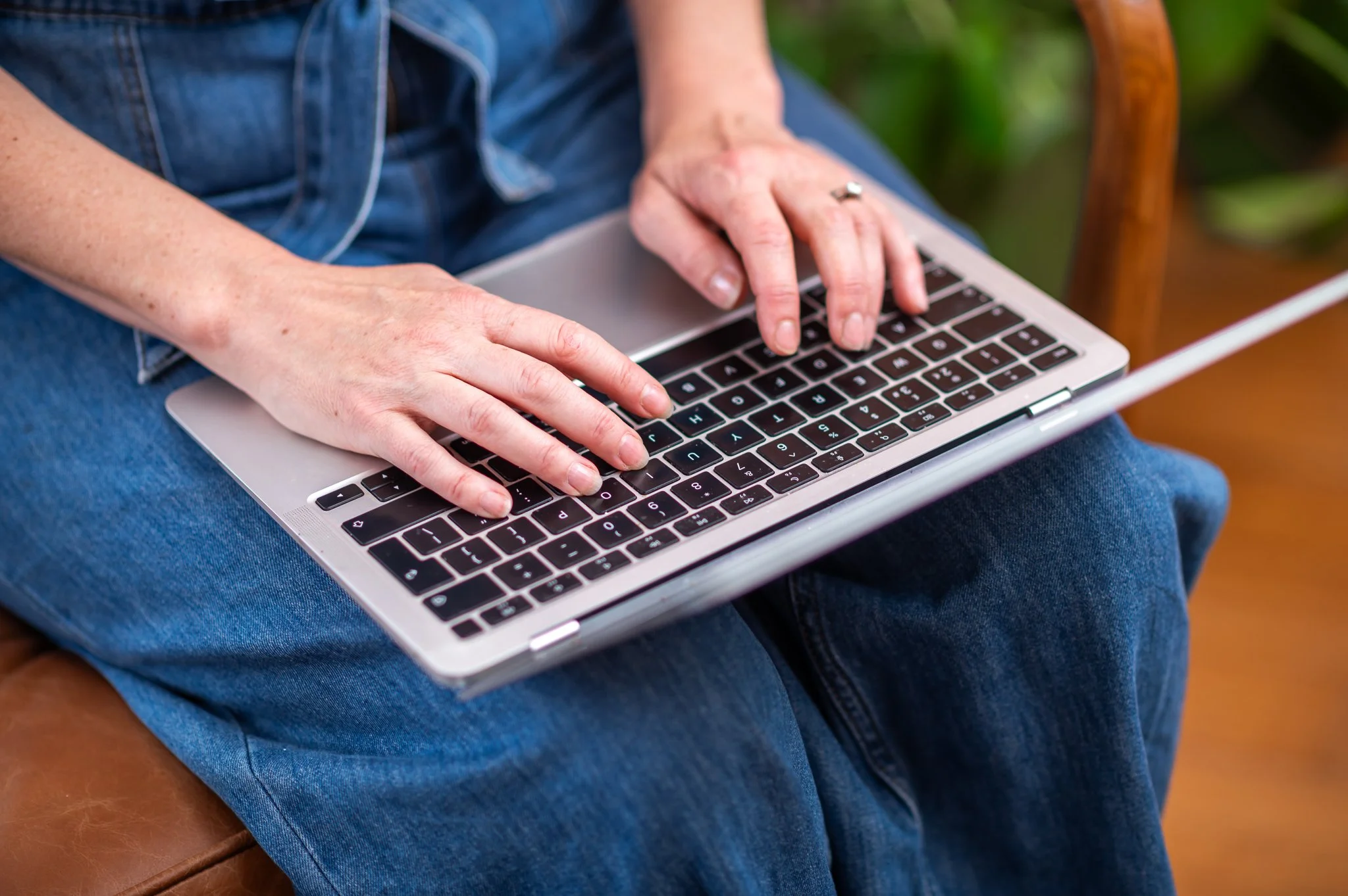 Person typing on a silver laptop with black keyboard, sitting on a brown leather chair, wearing blue jeans and a denim jacket.