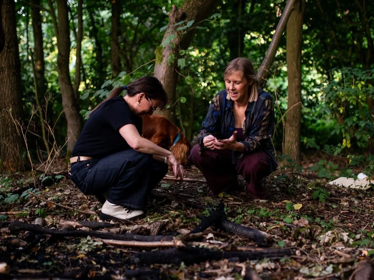Two women and a dog in a wooded forest, engaging with each other.