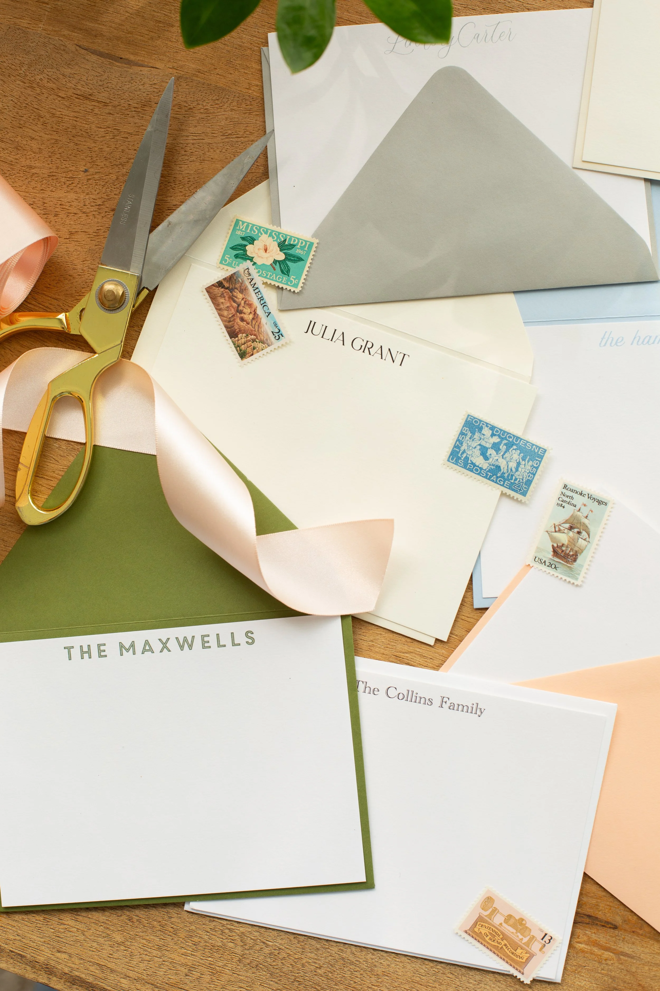Personalized stationery set with custom names on cards and envelopes, including “The Maxwells” and “Julia Grant,” styled with ribbon, scissors, and vintage stamps on a wooden desk.