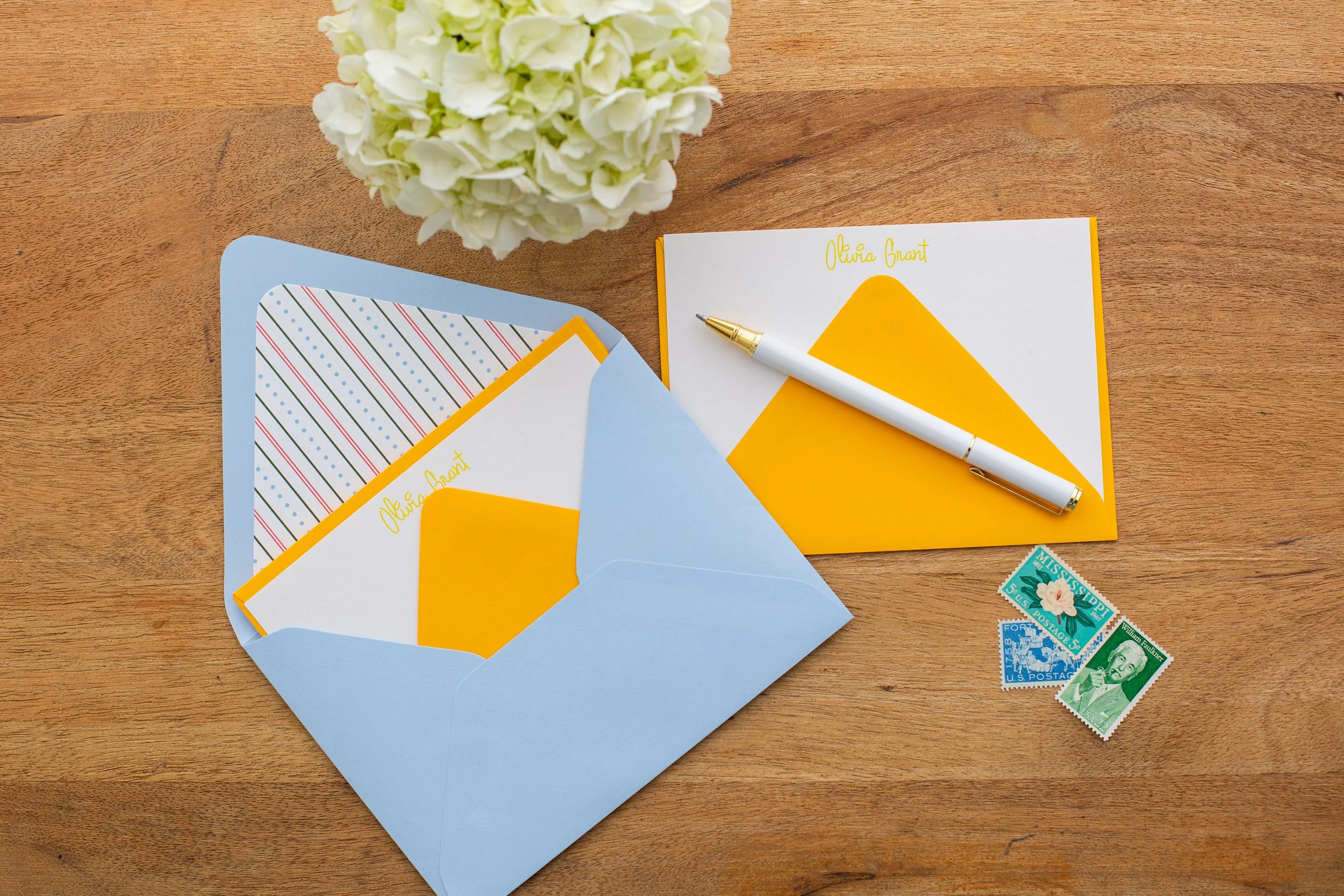 Personalized stationery set with geometric yellow designs and the custom name “Olivia Grant,” styled with blue envelopes, a white pen, and patterned paper on a wooden desk.