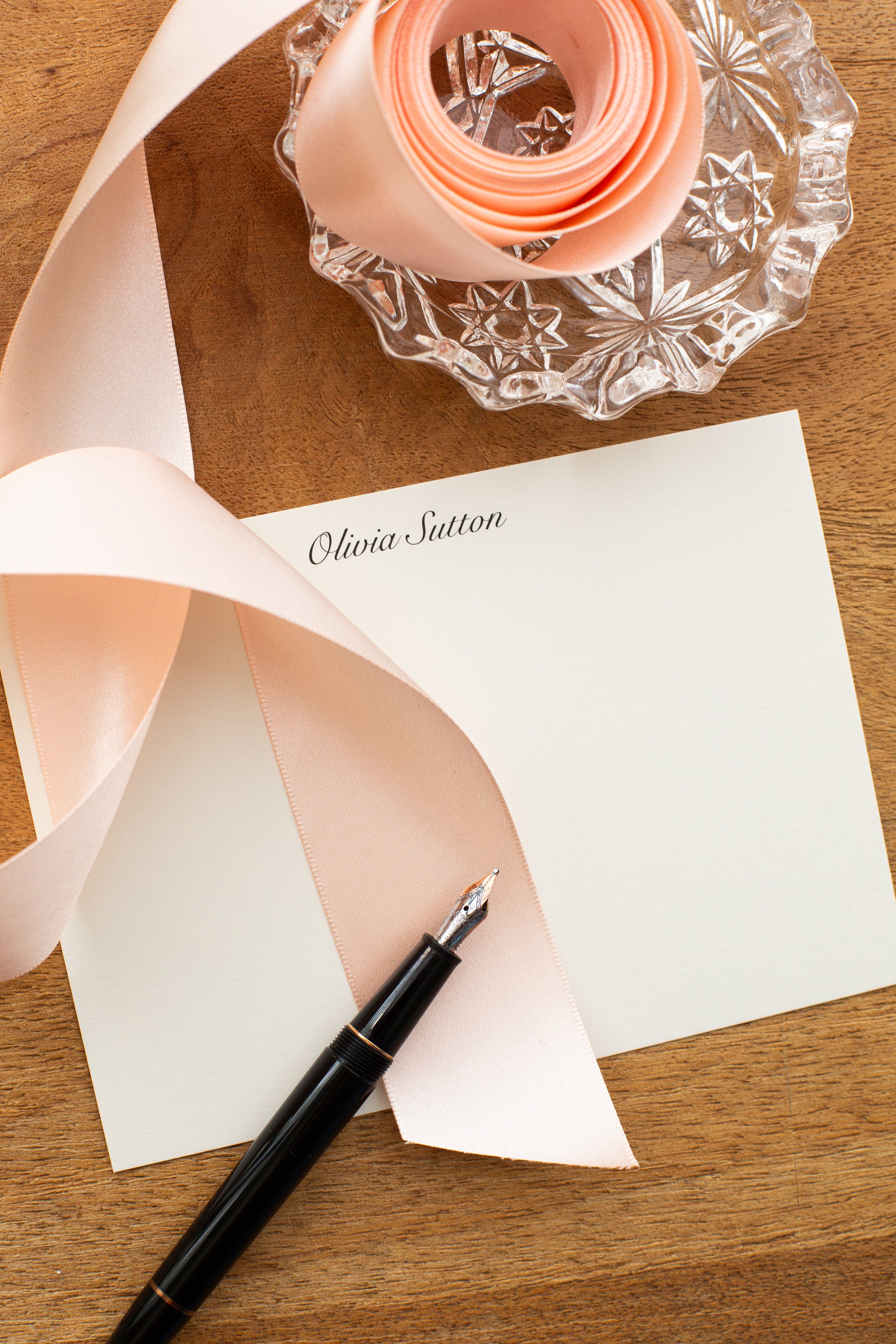 Close-up of personalized stationery with the name “Olivia Sutton,” styled with a blush ribbon, glass dish, and a black fountain pen on a wooden desk.