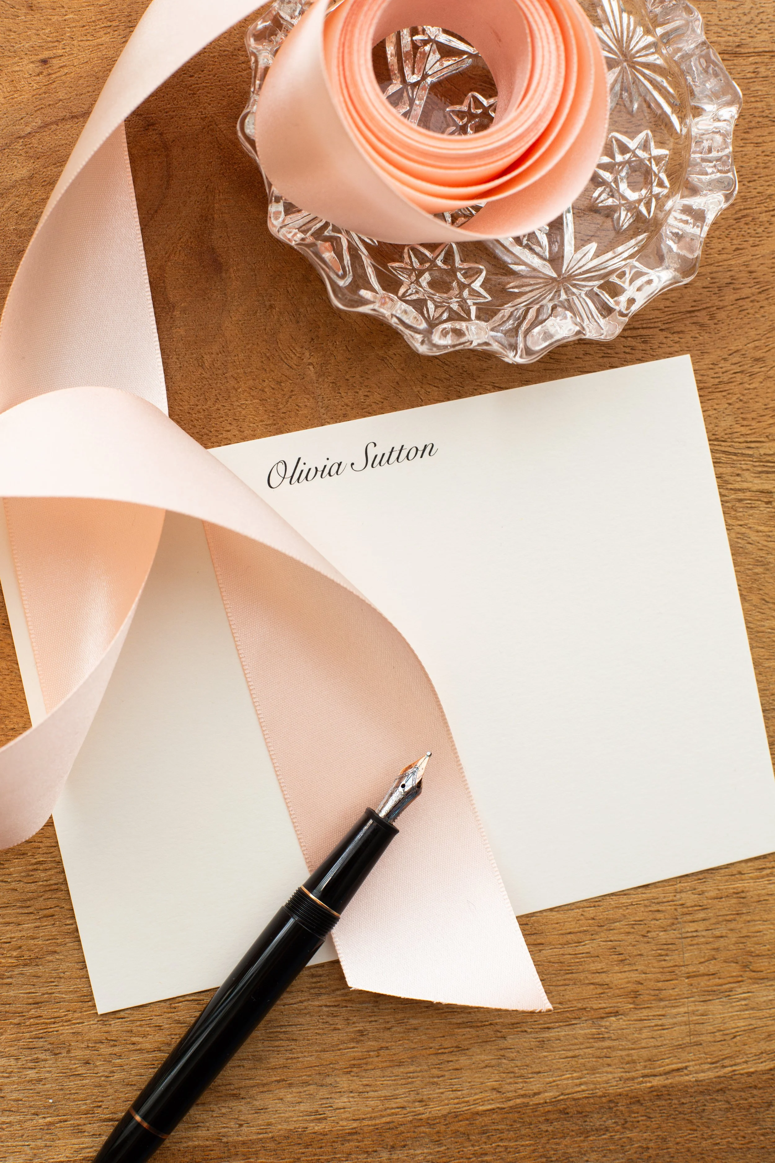 Close-up of personalized stationery with the name “Olivia Sutton,” styled with a blush ribbon, glass dish, and a black fountain pen on a wooden desk.