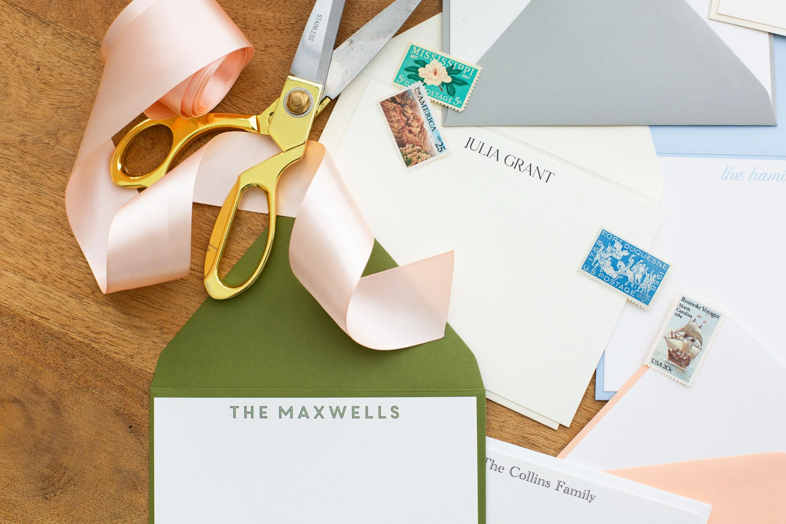 Personalized stationery set with custom names on cards and envelopes, including “The Maxwells” and “Julia Grant,” styled with ribbon, scissors, and vintage stamps on a wooden desk.