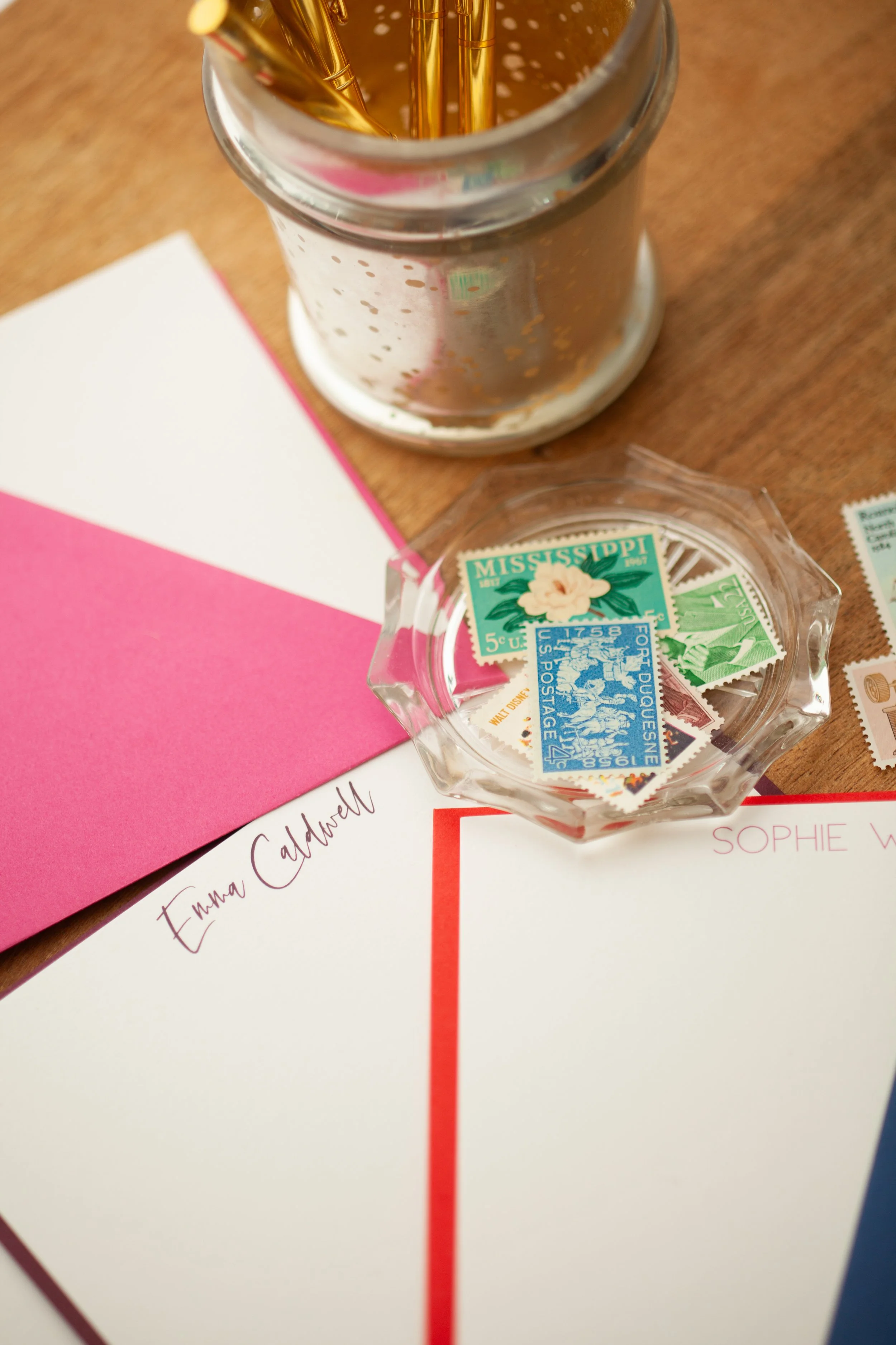 Personalized stationery cards with colorful borders and custom names, including “Emma Caldwell,” styled with vintage stamps and a glass dish on a wooden desk.