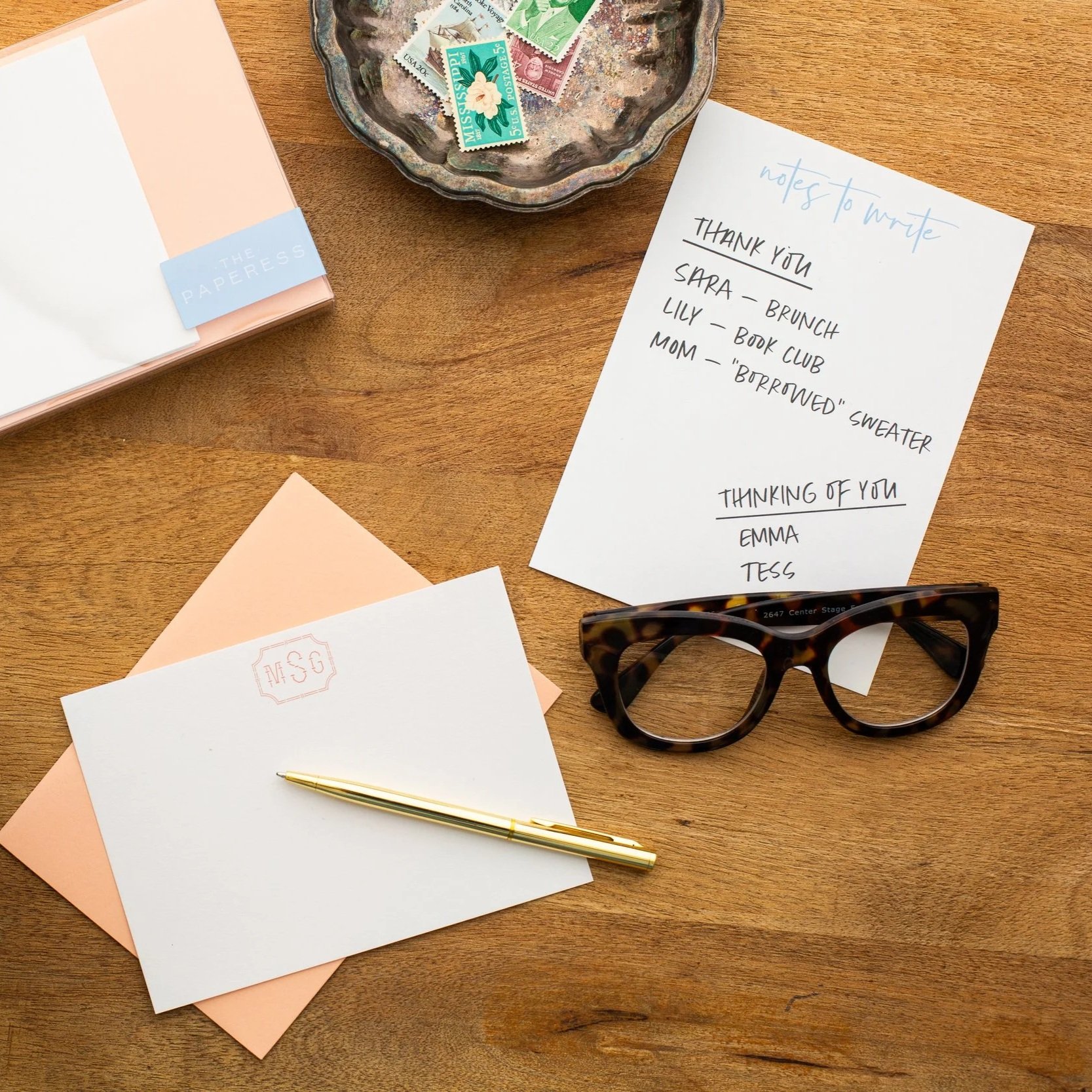 Flat lay of personalized stationery, envelopes, and a gold pen on a wooden desk beside tortoiseshell glasses and a handwritten note listing reminders to write thank you and thinking-of-you cards.