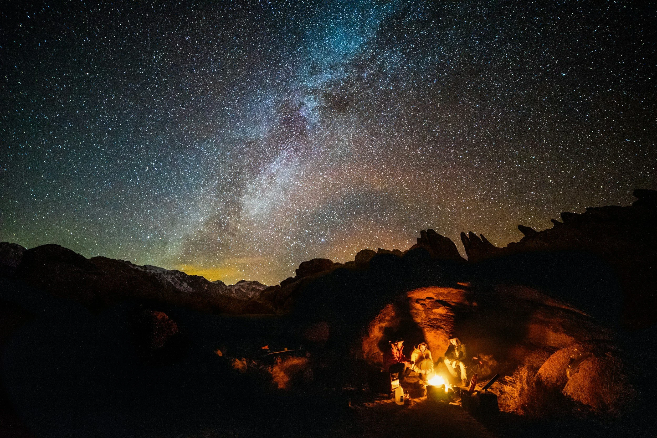 Night sky filled with countless stars and the Milky Way galaxy, overlooking a rocky mountain landscape with campfire on the ground illuminating a small group of people.