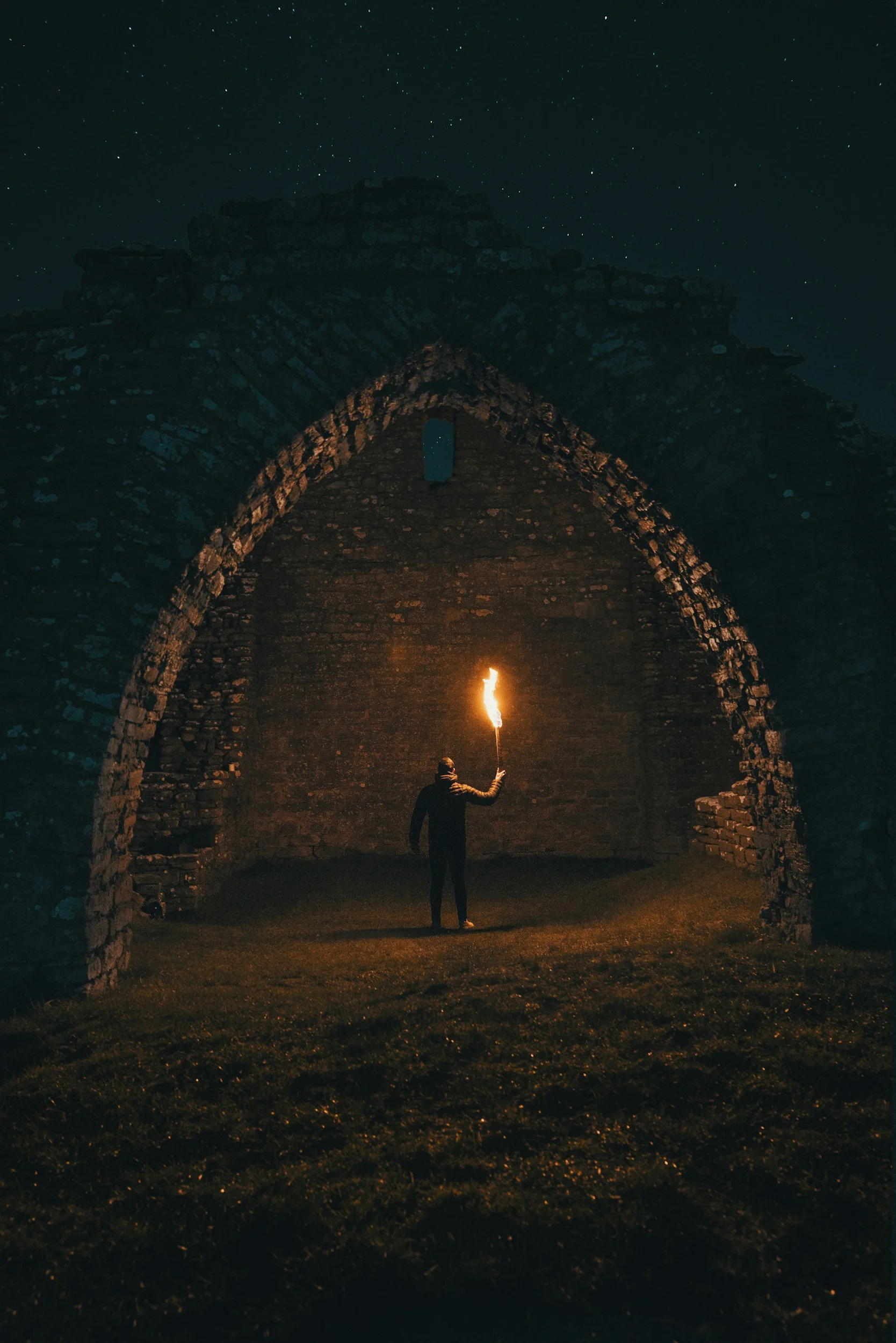 A person holding a flaming torch standing underneath a large, ancient stone archway at night, with a starry sky overhead.