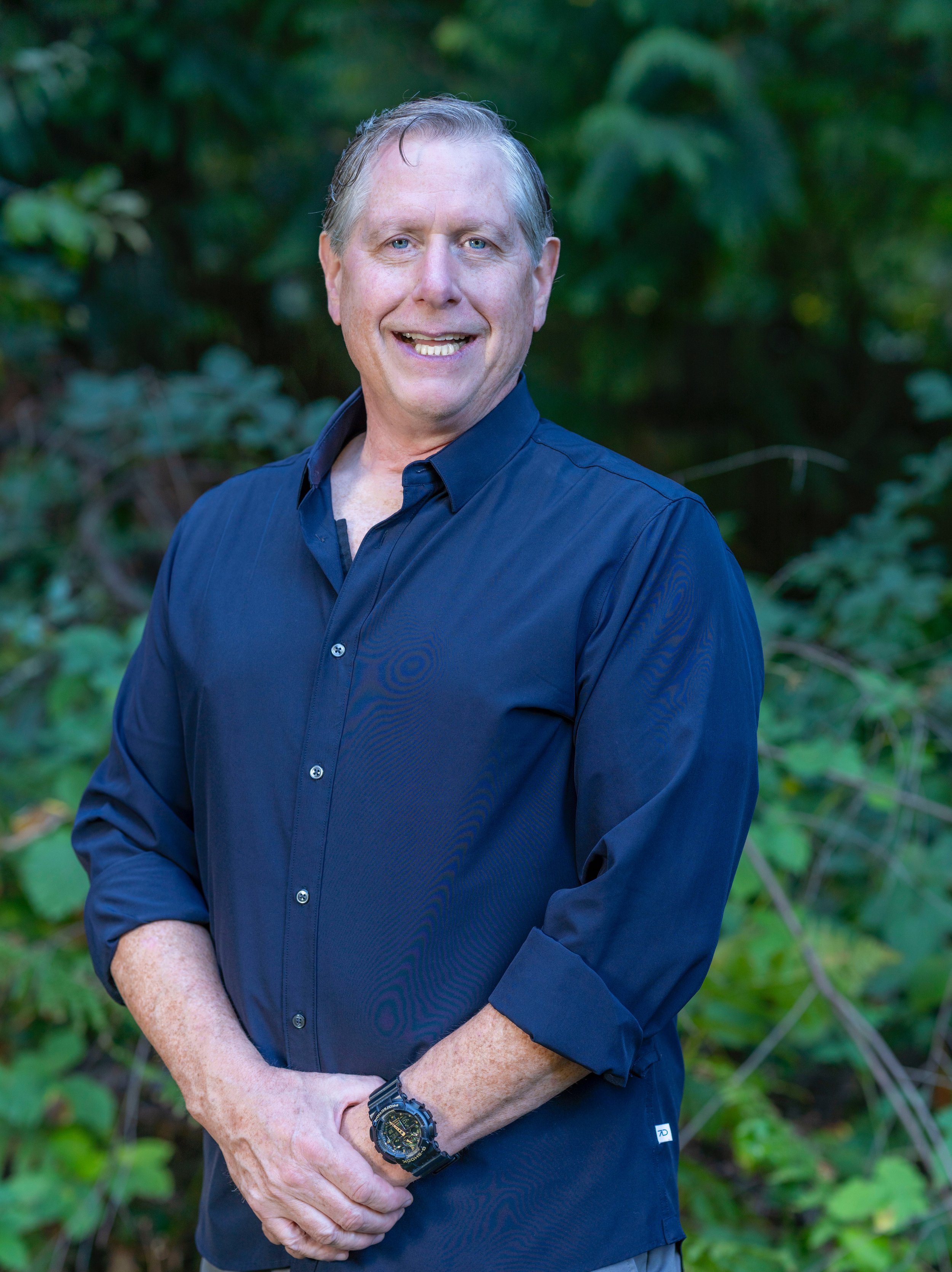 A middle-aged man with light hair and blue eyes smiling in front of a green, leafy background, wearing a dark blue button-up shirt and a black watch.