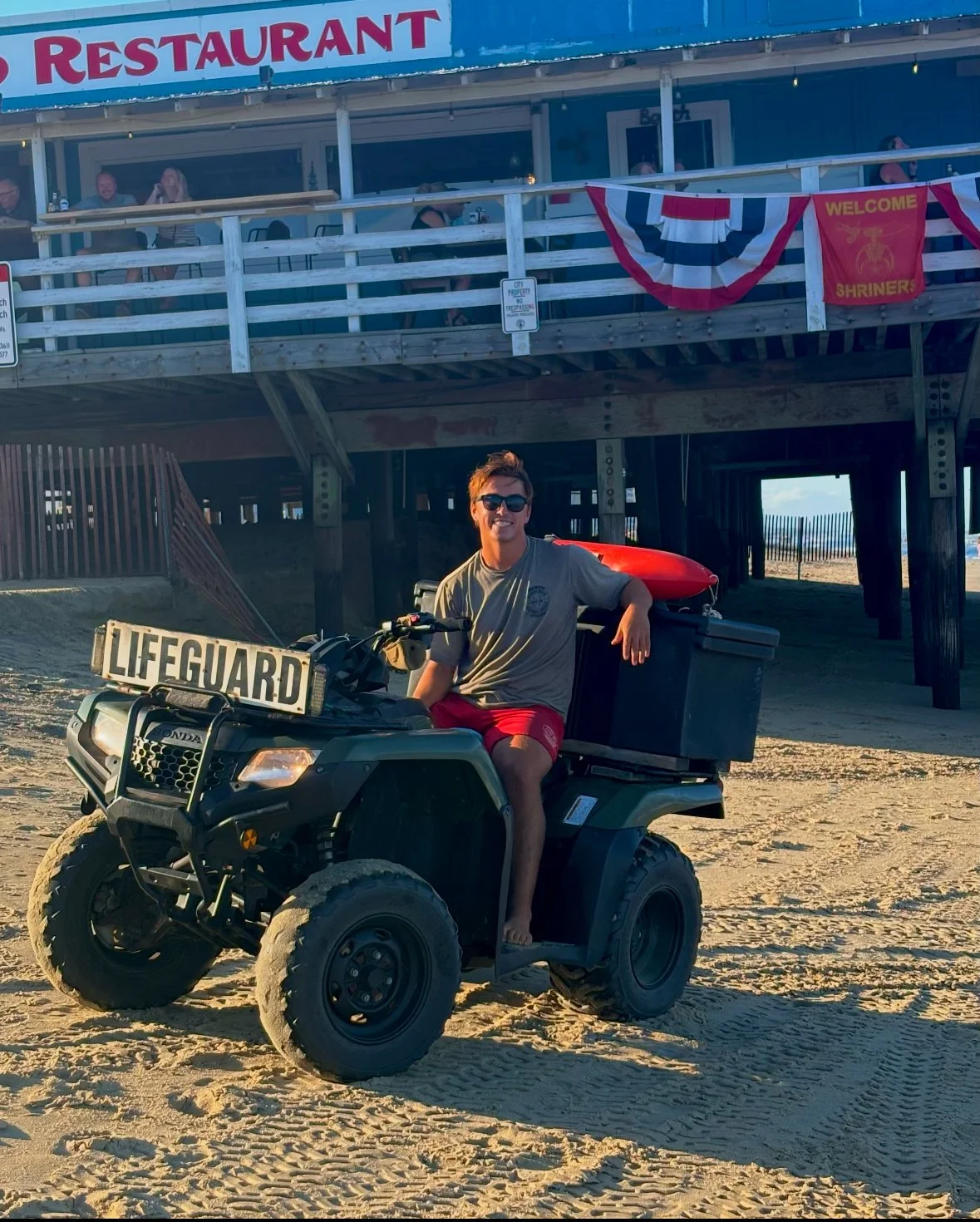 Young man wearing sunglasses, sitting on an all-terrain vehicle with a 'LIFEGUARD' sign, at the beach near a pier with a restaurant and people on the upper deck.