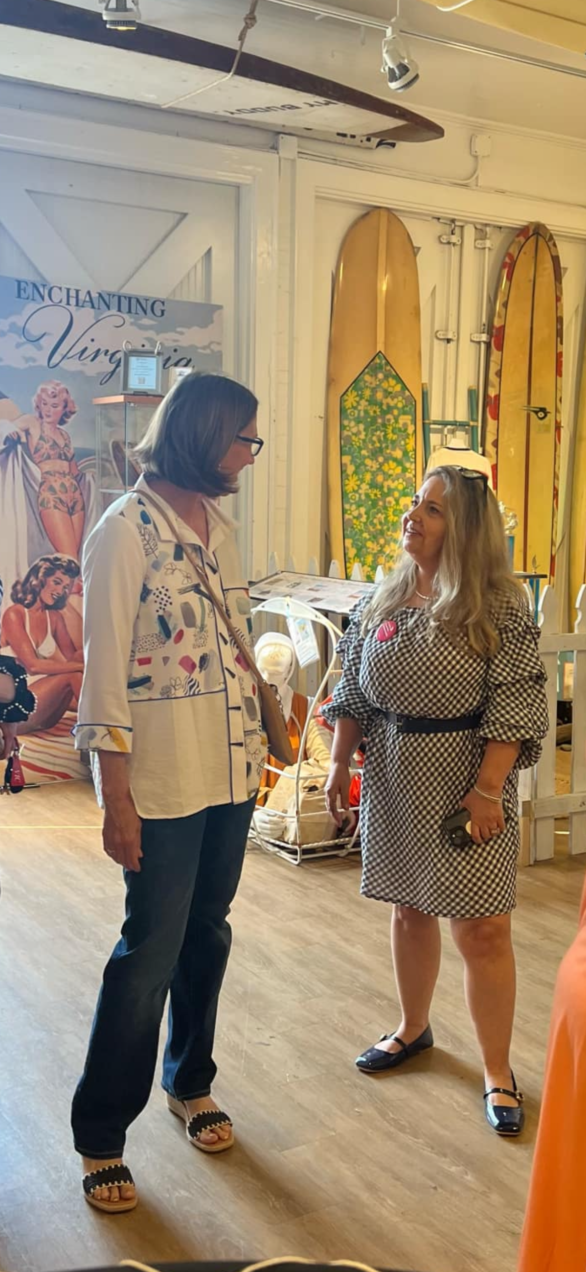 Two women are engaged in a conversation inside a vintage shop with surfboards and vintage posters in the background.