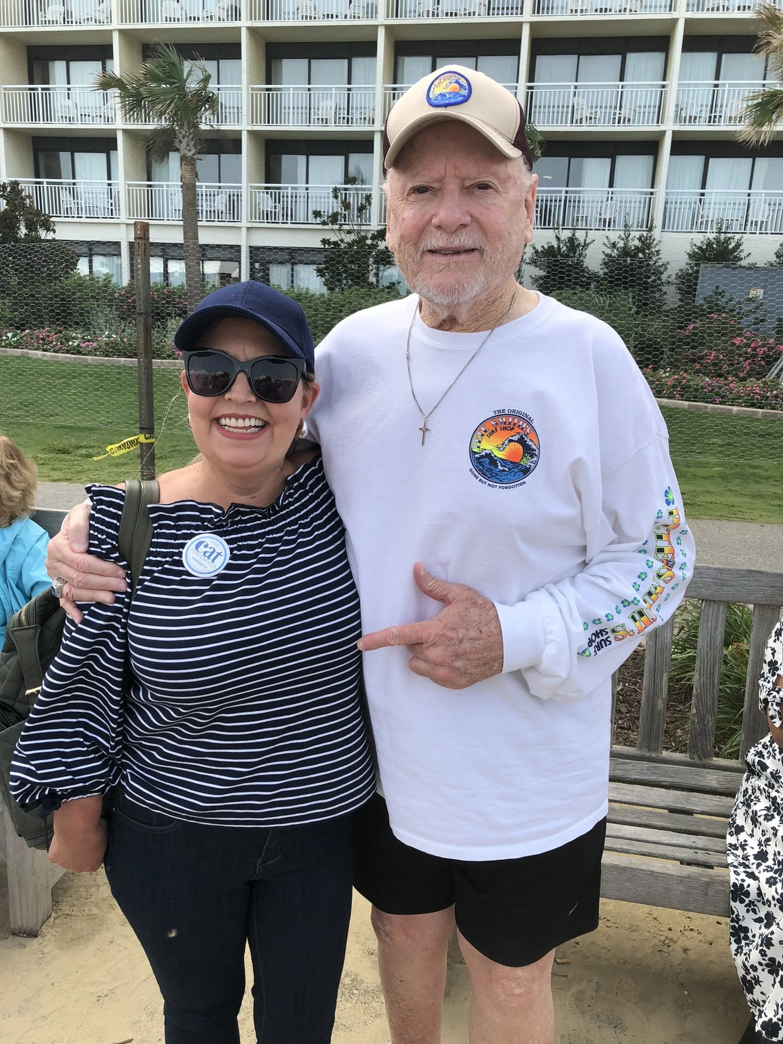 A smiling woman in sunglasses and a navy blue hat standing next to a man in a tan hat and white long sleeve shirt, pointing at him, outdoors with a building with balconies and palm trees in the background.