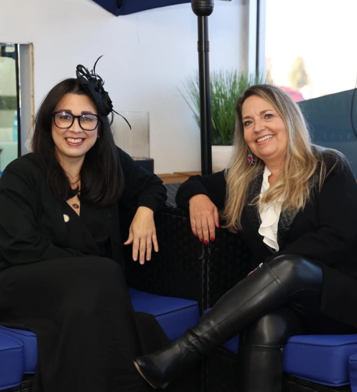 Two women sitting on blue cushions outside, smiling at the camera. One woman has long dark hair, glasses, and a decorative black hat. The other woman has long blonde hair and colorful earrings. They are sitting near a large plant and umbrella.