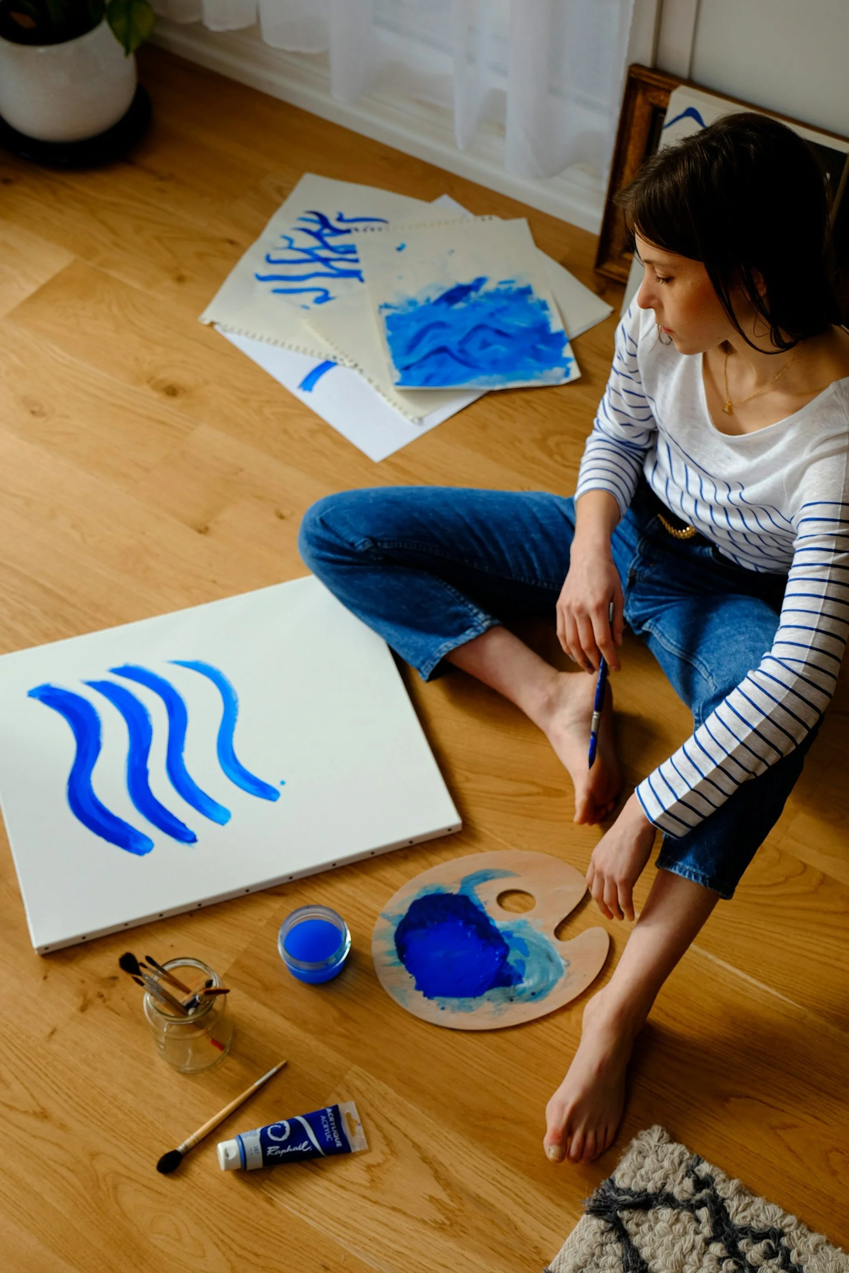 someone sat on a wood floor painting blue wavy lines on a canvas.