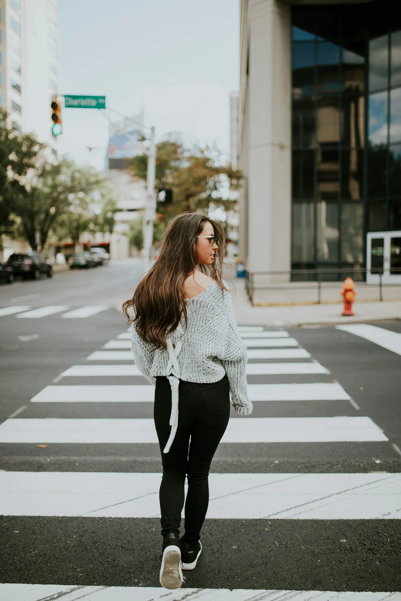 a woman, wearing a jumper, leggings, and trainers, is crossing the road in a city. She is looking over her shoulder and has long brown hair