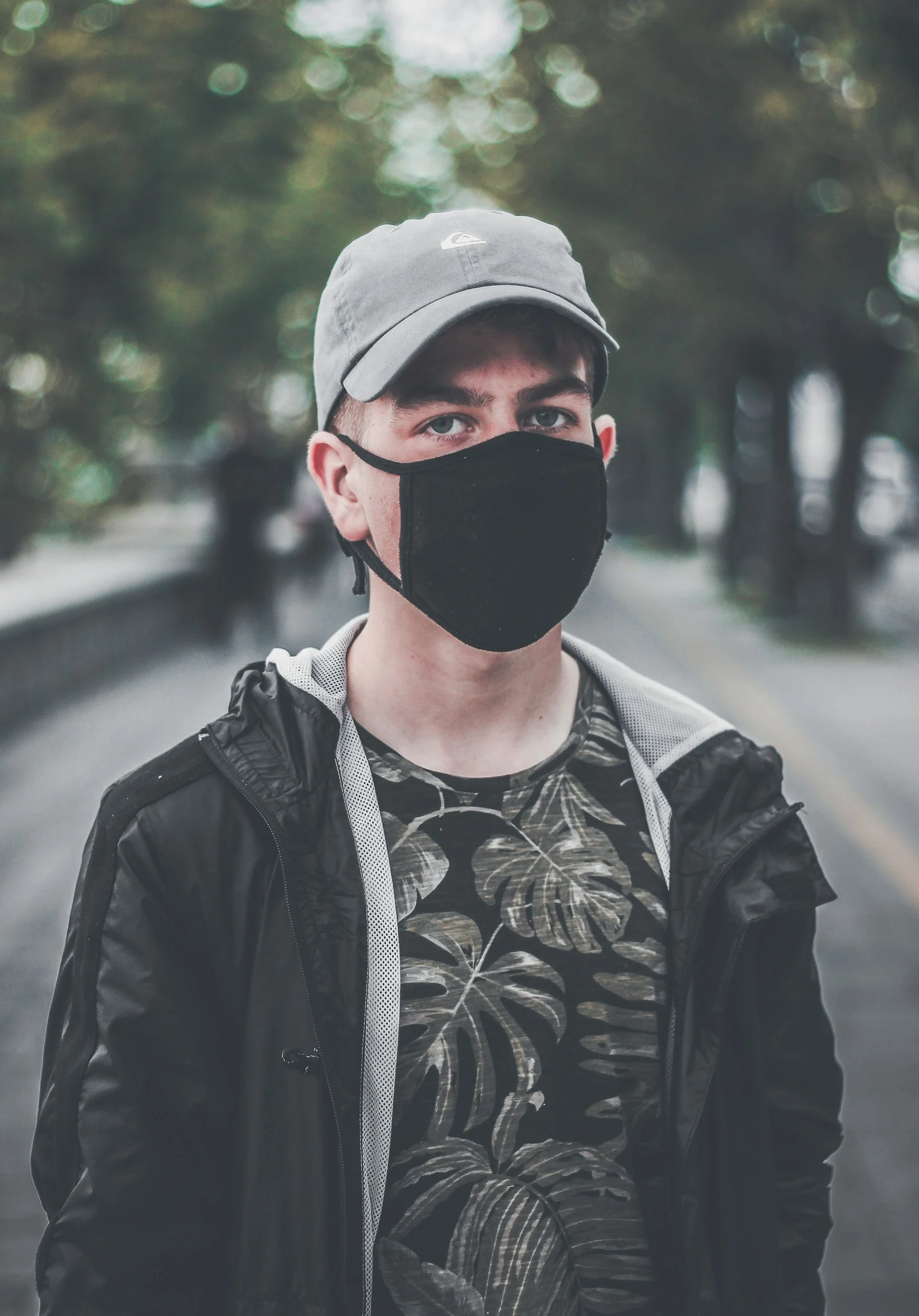an image of a person with a black face mask and a grey hat. They have a neutral look on their face and are wearing a black jacket and a black shirt with green monstera leaves on it.