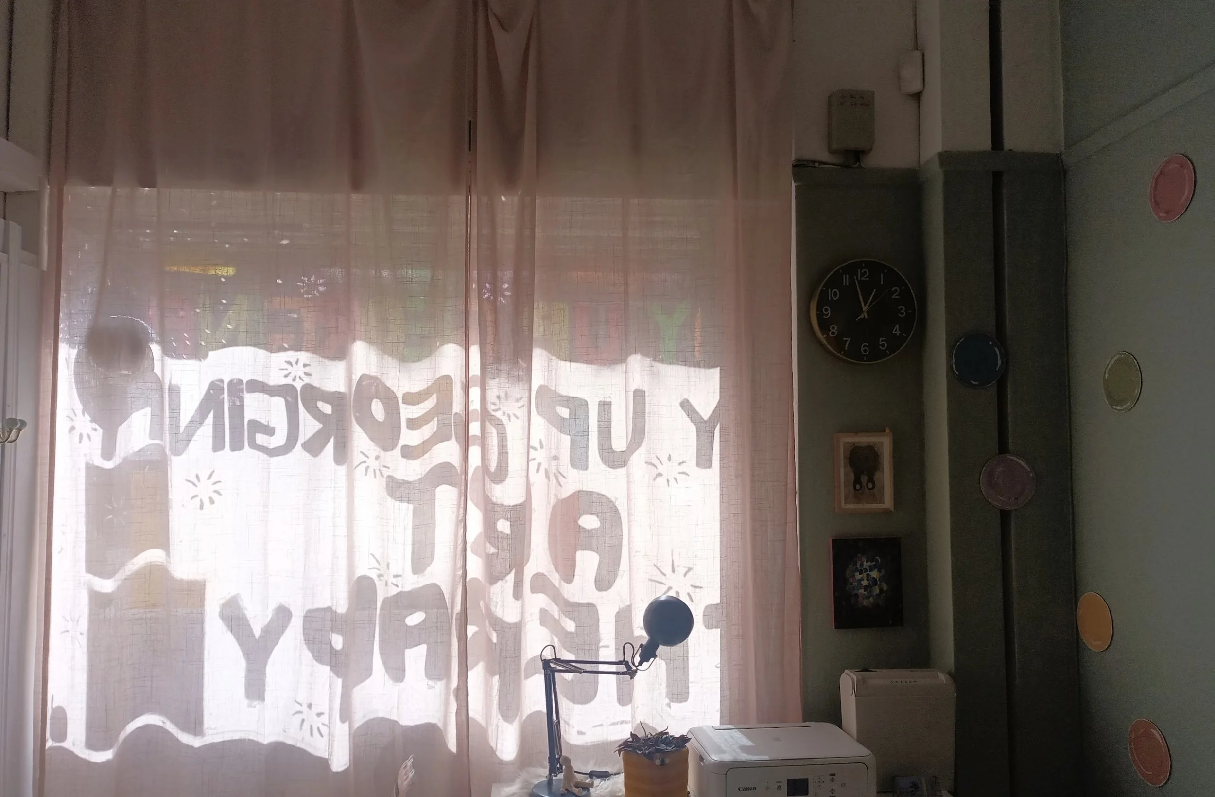 an image of the art therapy studio window behind pale pink curtains. You can see the shadow of the words “Ay up Georgina art therapy” from the sun shining through the window display.