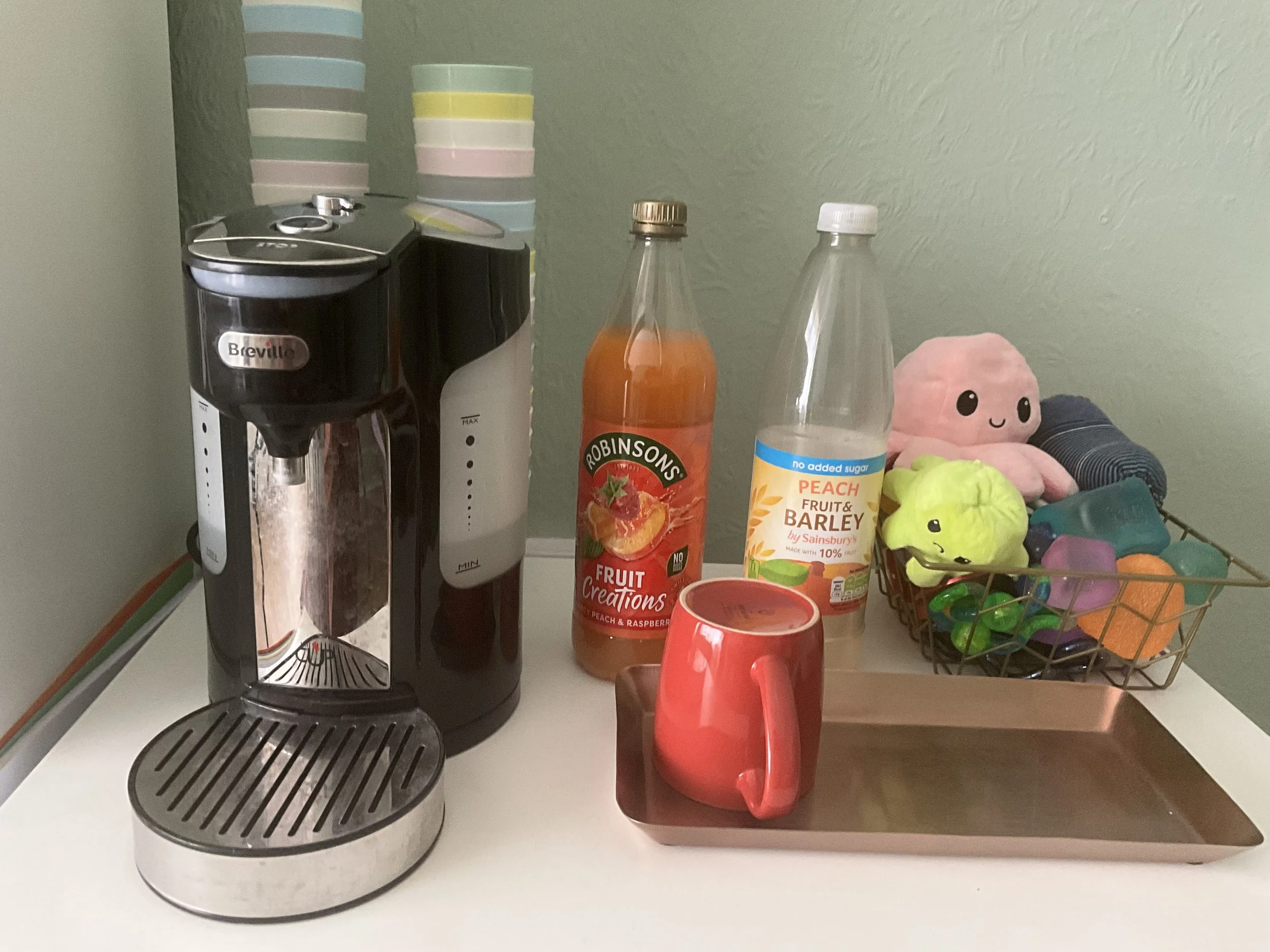 a picture of a kettle that brews one cup, a rose gold tray with a mug on it, bottles of peach and raspberry squash, and a bowl of fidgets