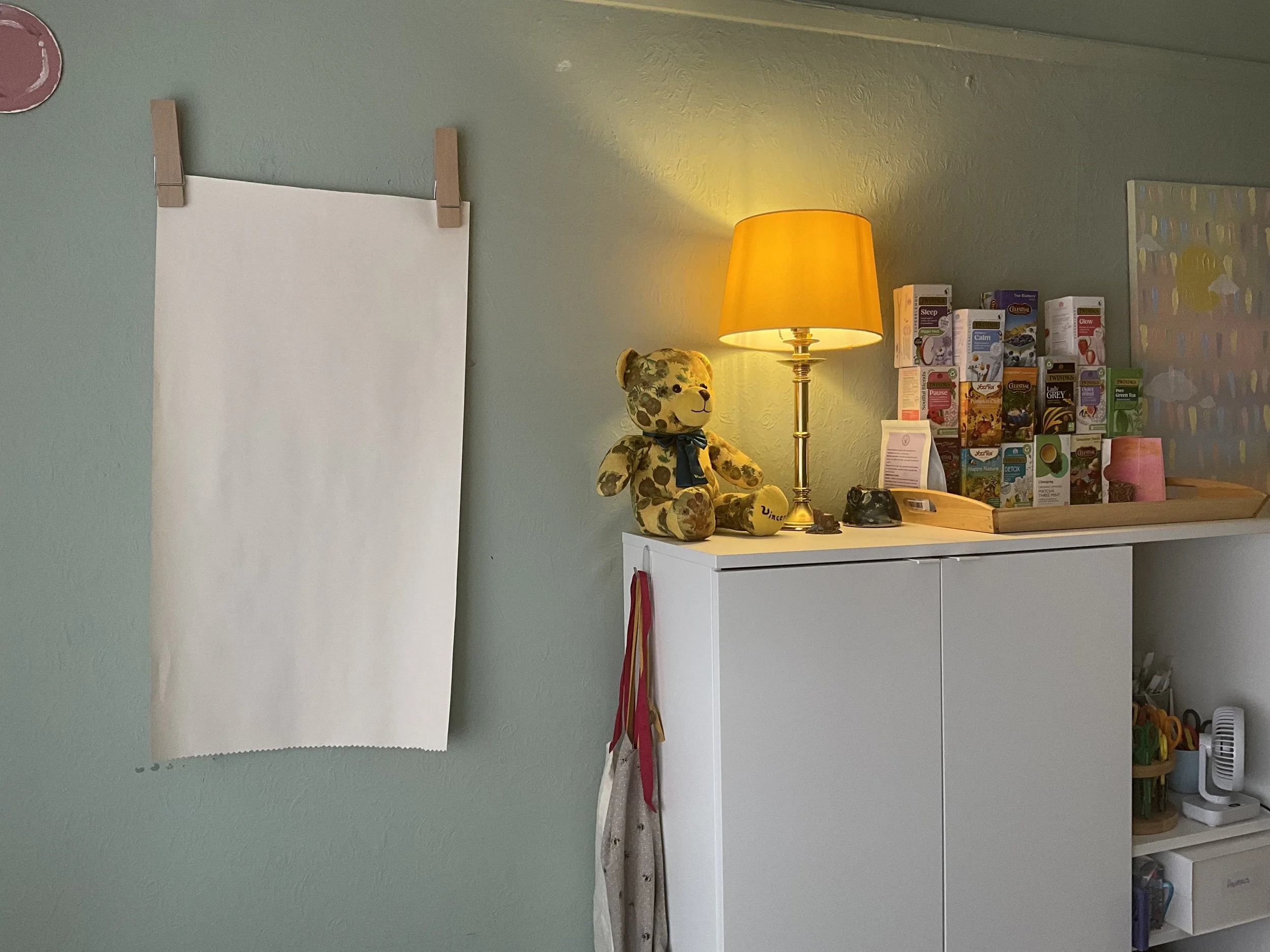 Two large wooden pegs holding up a large piece of paper on the sage green wall, to the left of the materials cupboard in the therapy space
