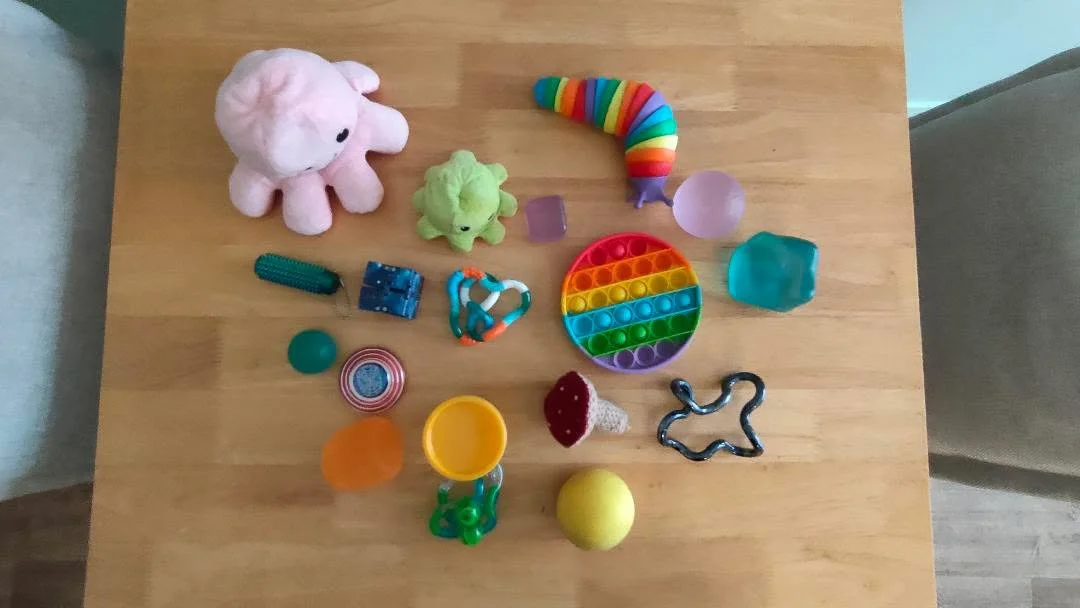 A selection of fidgets on the therapy studio table. Fidgets include various squish balls and cubes, tangles, playdoh, slugs, pop fidgets, cube fidgets, and octopus fidgets