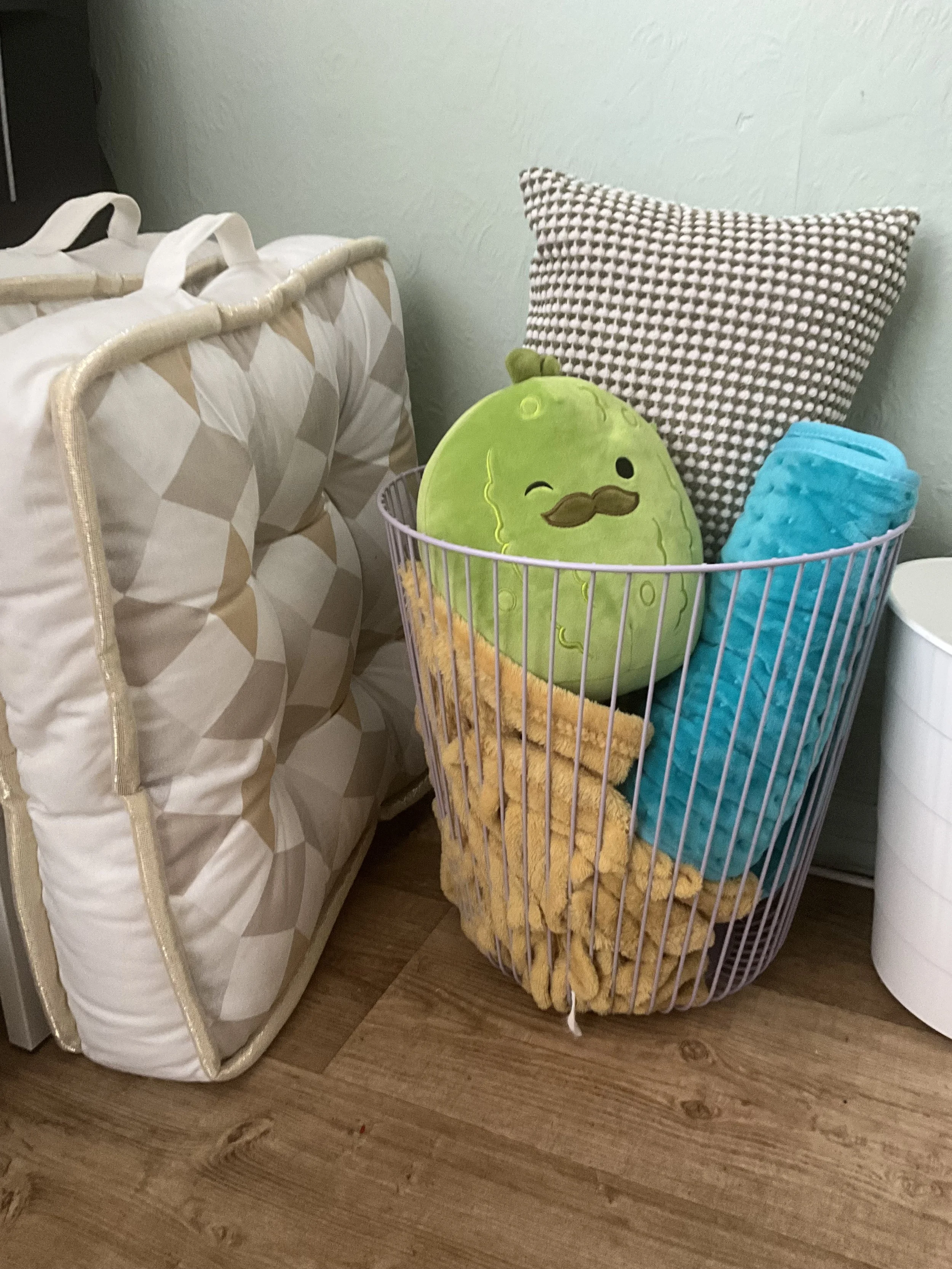 A lilac wire basket with a yellow fuzzy blanket, a blue electric heat pad, a green pickle squishmallow, and a green and white cushion. Next to a recyling bin and floor cushions.