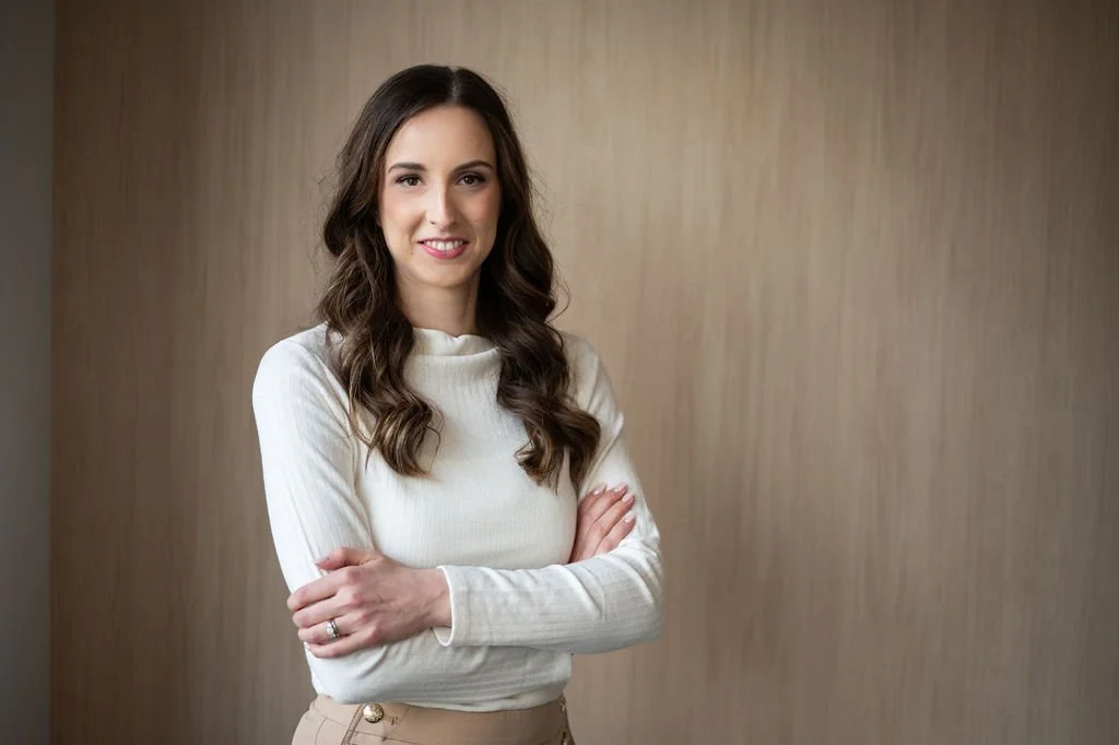 A young woman with long brown wavy hair, wearing a cream-colored long-sleeve top and beige pants, standing with her arms crossed and smiling at the camera against a plain, slightly textured beige background.