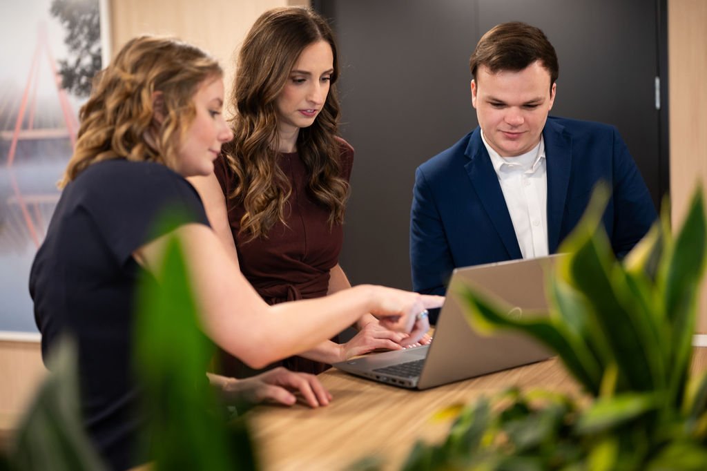 Three young professionals, two women and one man, are gathered around a laptop at a meeting table, collaborating and discussing.