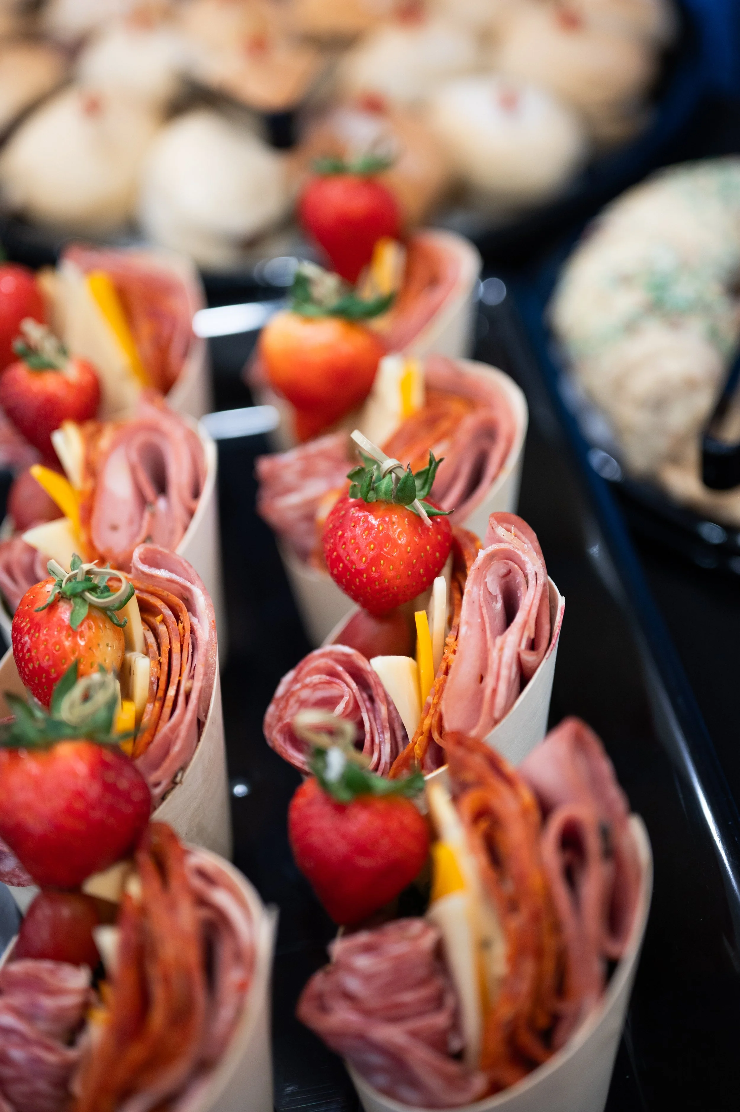 Mini sandwiches topped with strawberries and cheese, arranged on a tray, with assorted other finger foods in the background.